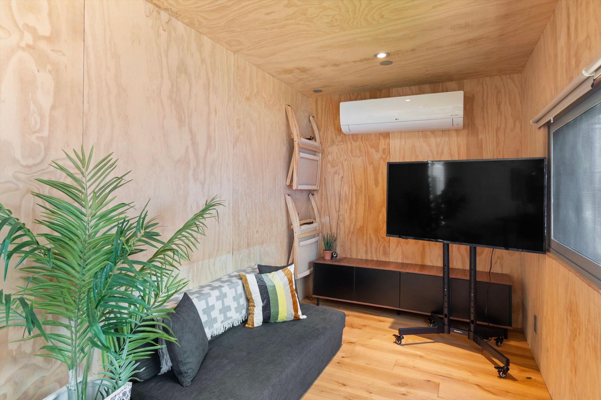 A small, wood-paneled room with a couch, TV on a rolling stand, air conditioner, and a ladder shelf. A large potted plant sits near the couch.