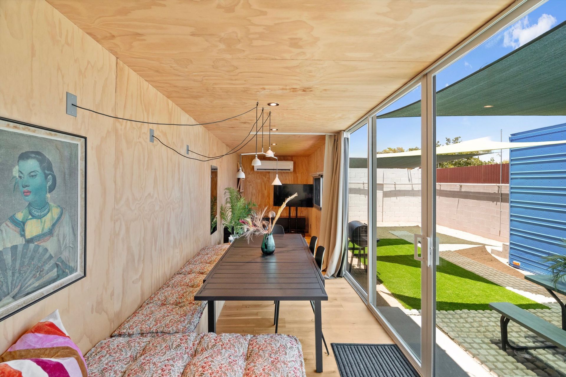 Interior of a long, narrow room with a dining table and bench seating, a large window overlooking a yard, and artwork on the wall.