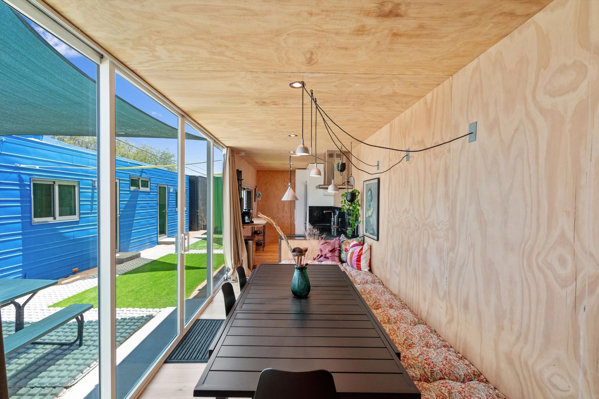 Long dining table in a container home with a built-in bench seat, large windows, and a view of a blue building and yard.