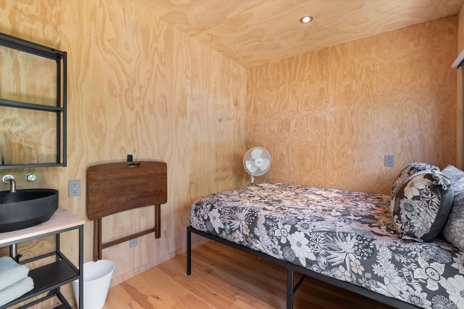 A small, wood-paneled bedroom with a single bed, wall-mounted sink, and a fold-down table.