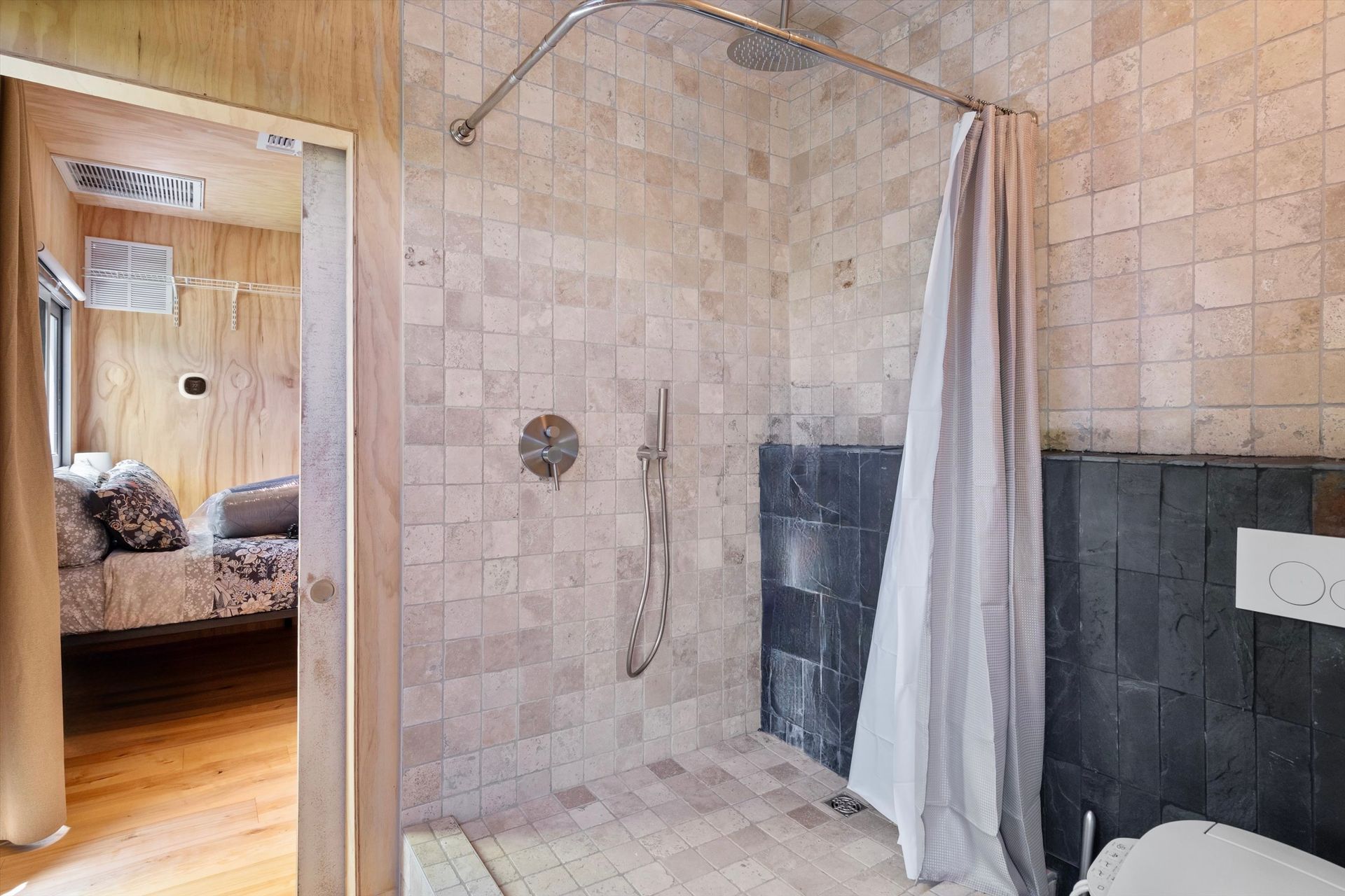 Bathroom with a shower, wood paneling, and a view into a bedroom. Shower features stone tiles and a gray shower curtain.