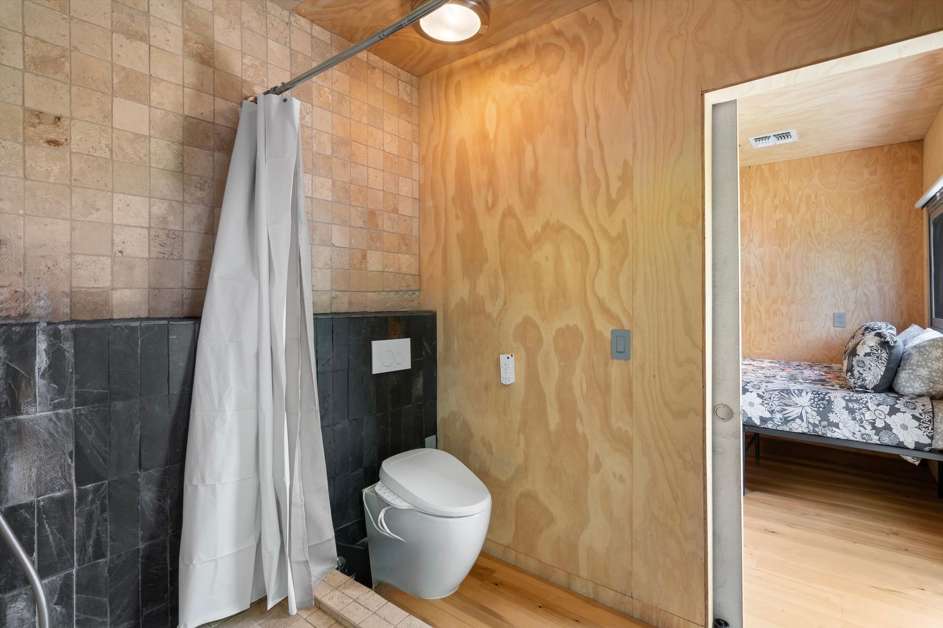Small bathroom with a toilet, shower curtain, and plywood walls. A bedroom is visible through an open doorway.
