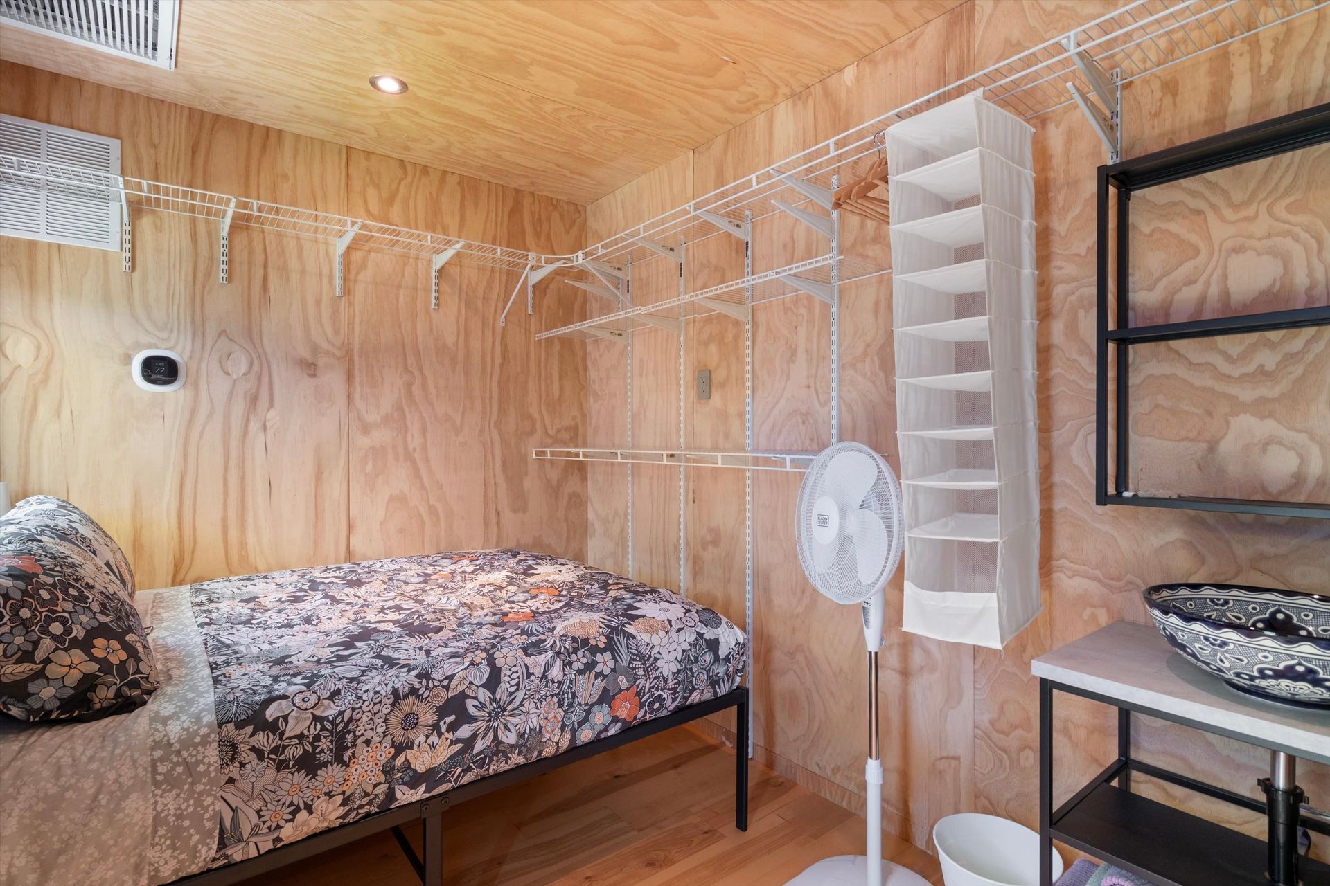 Small, rustic bedroom with wooden walls, a bed, hanging shelves, a fan, and a sink.
