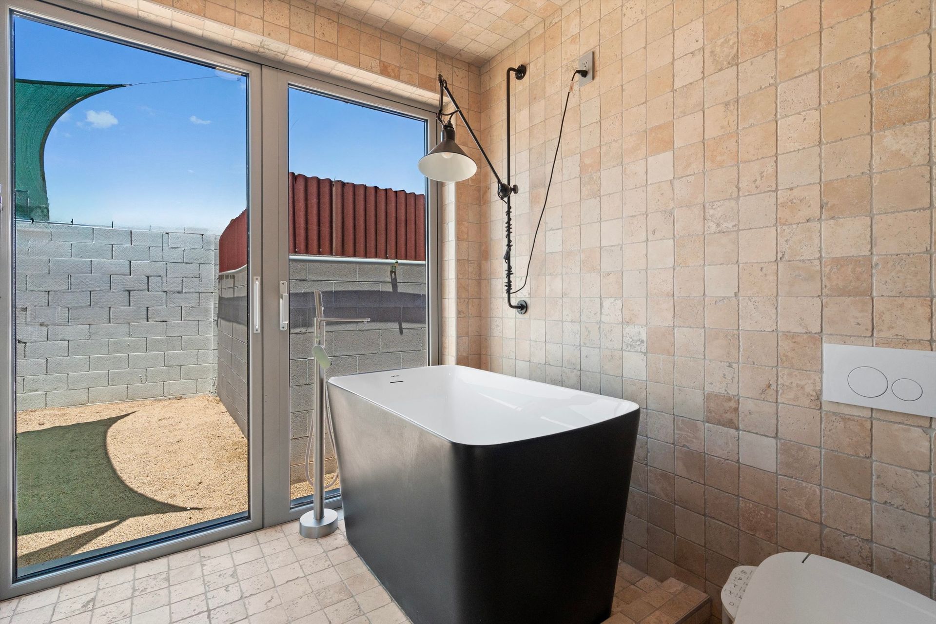 A modern bathroom with a black bathtub next to a large window overlooking a patio. The walls are tiled, and there is a shower head.