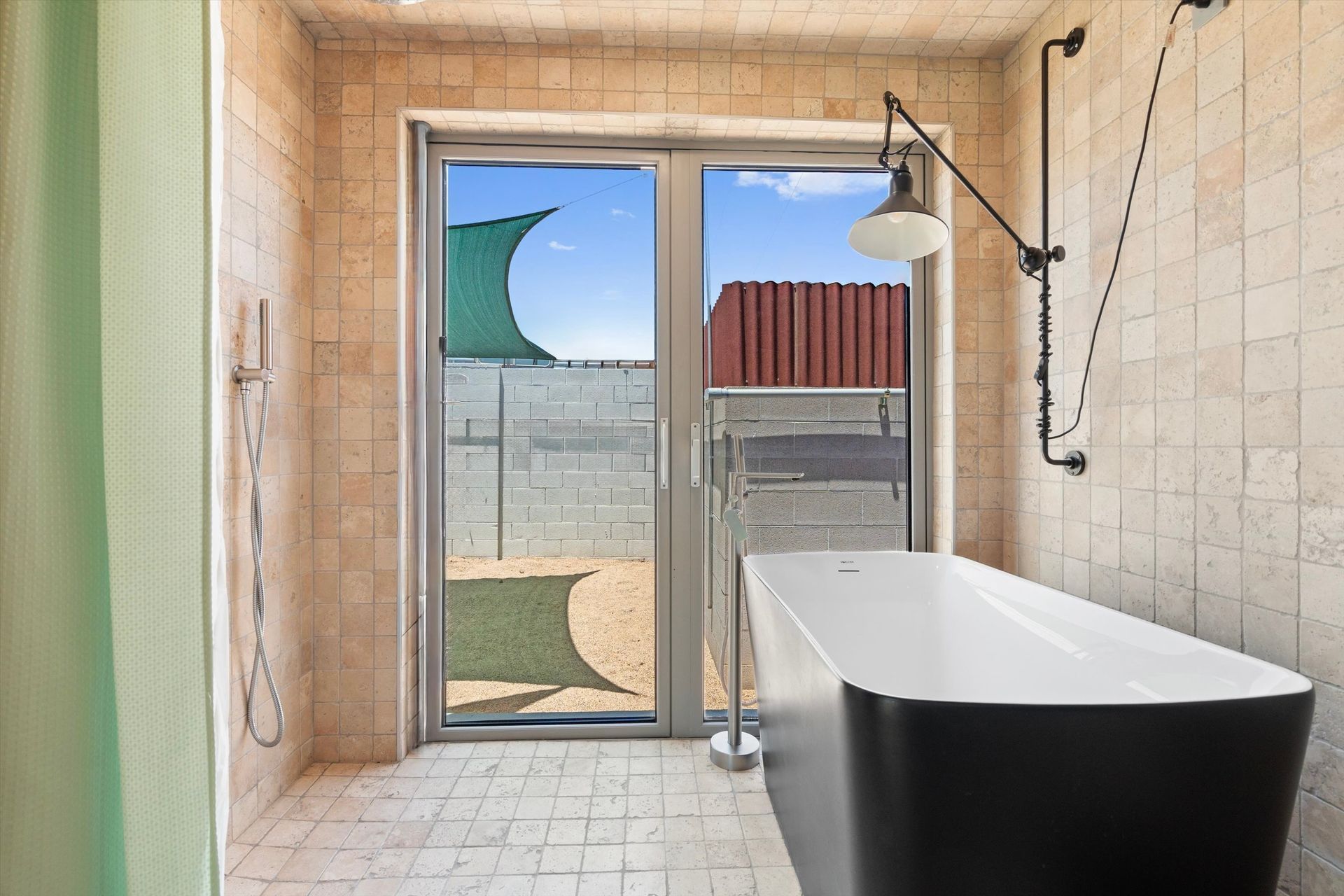 Modern bathroom with a black freestanding bathtub, open to a small outdoor patio. Beige tiled walls, a black industrial-style lamp, and a view of a fence are visible.