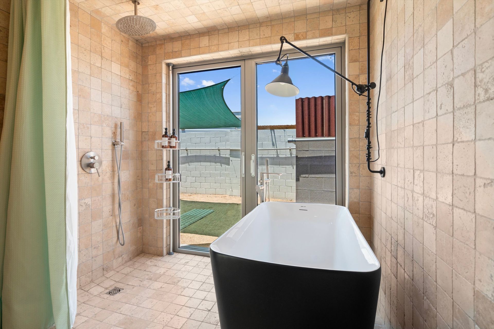 Bathroom with a black freestanding tub, shower, and a window view of colorful rooftops. Light tan tiles cover the walls.