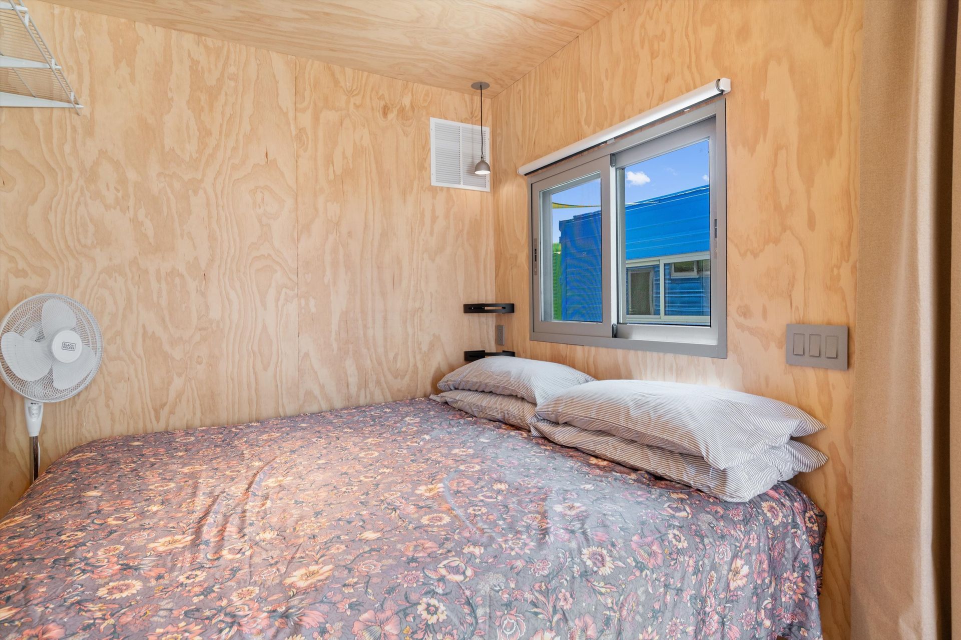 Small bedroom with wooden walls, a bed with patterned bedding, and a window with a blue view. A fan is to the left of the bed.