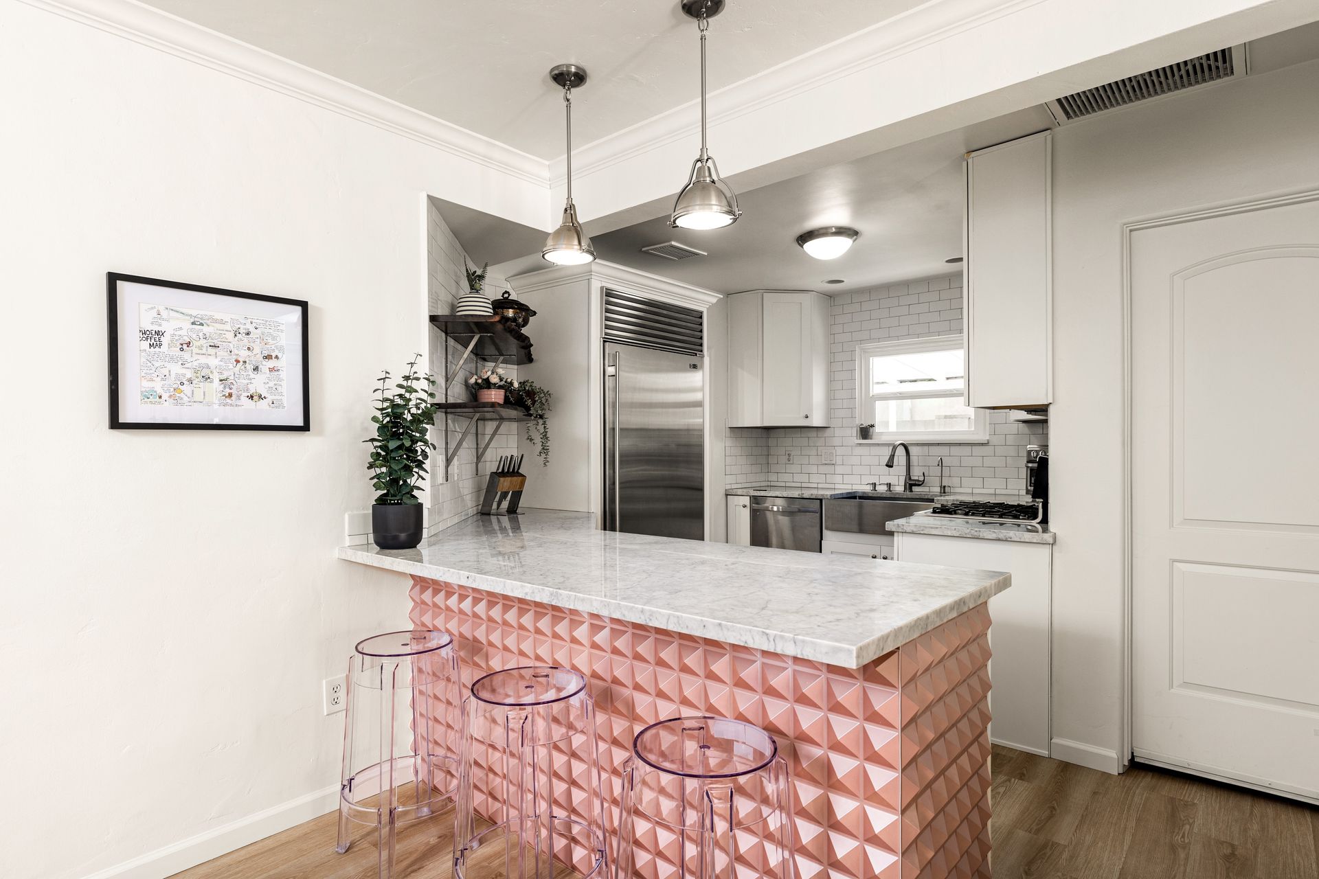 A modern kitchen with a pink tiled bar, a marble countertop, and clear acrylic bar stools. Overhead, two pendant lights illuminate the space.