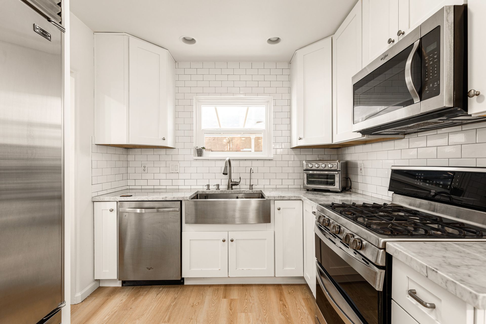 A modern white kitchen featuring stainless steel appliances, a farmhouse sink, and light wood floors. White subway tile backsplash.