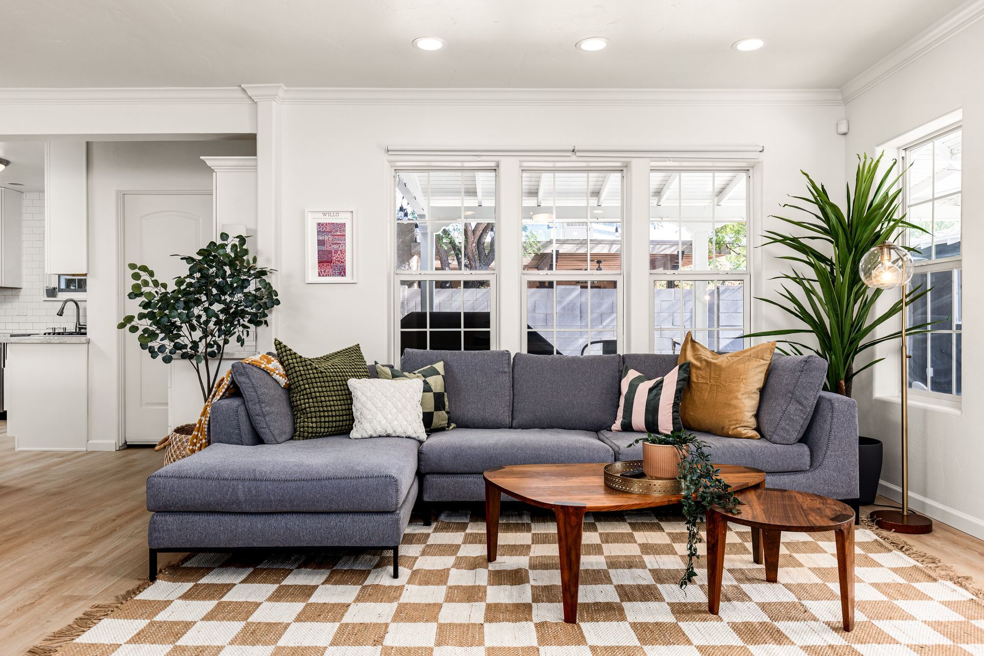 Cozy living room with a blue sectional sofa, wooden coffee tables, and a checkered rug. Large windows let in natural light.