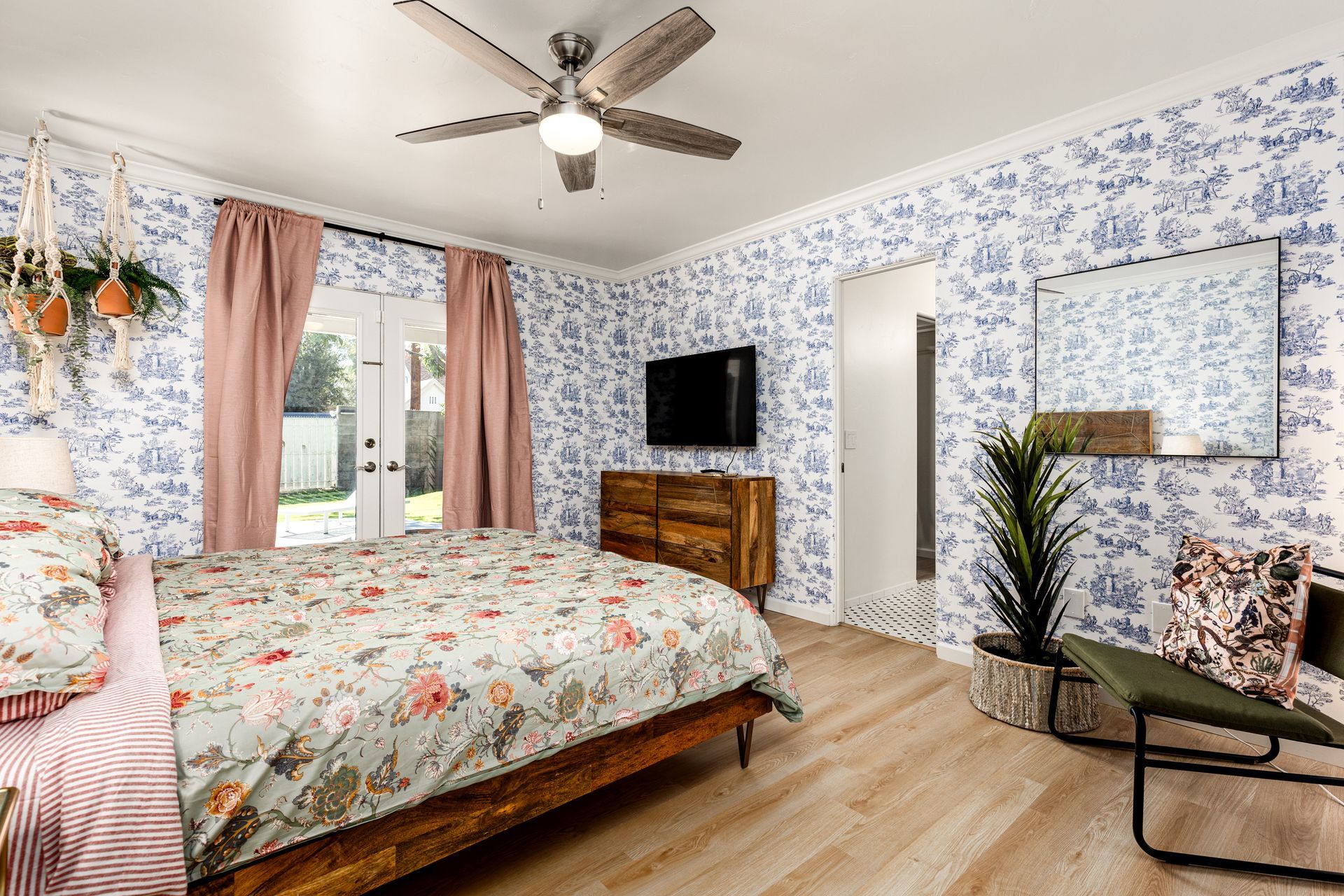 Bedroom with floral wallpaper, wooden furniture, pink curtains, and a large bed with patterned bedding. There's a door leading to a backyard.