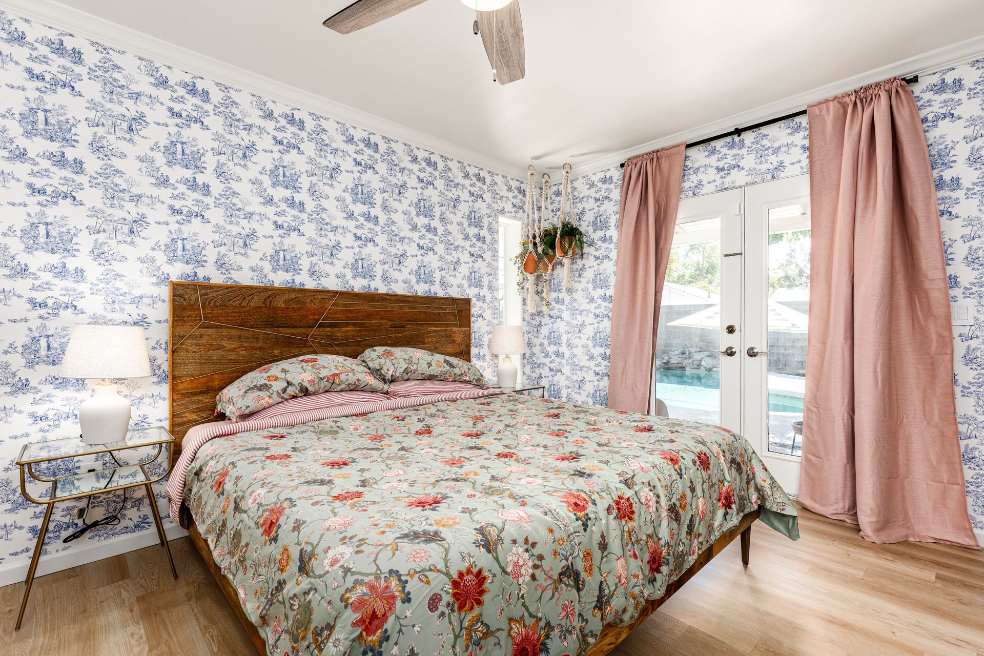 Bedroom with blue floral wallpaper, wood headboard, pink curtains, and a bed with floral bedding.  A small table with a lamp sits next to the bed.