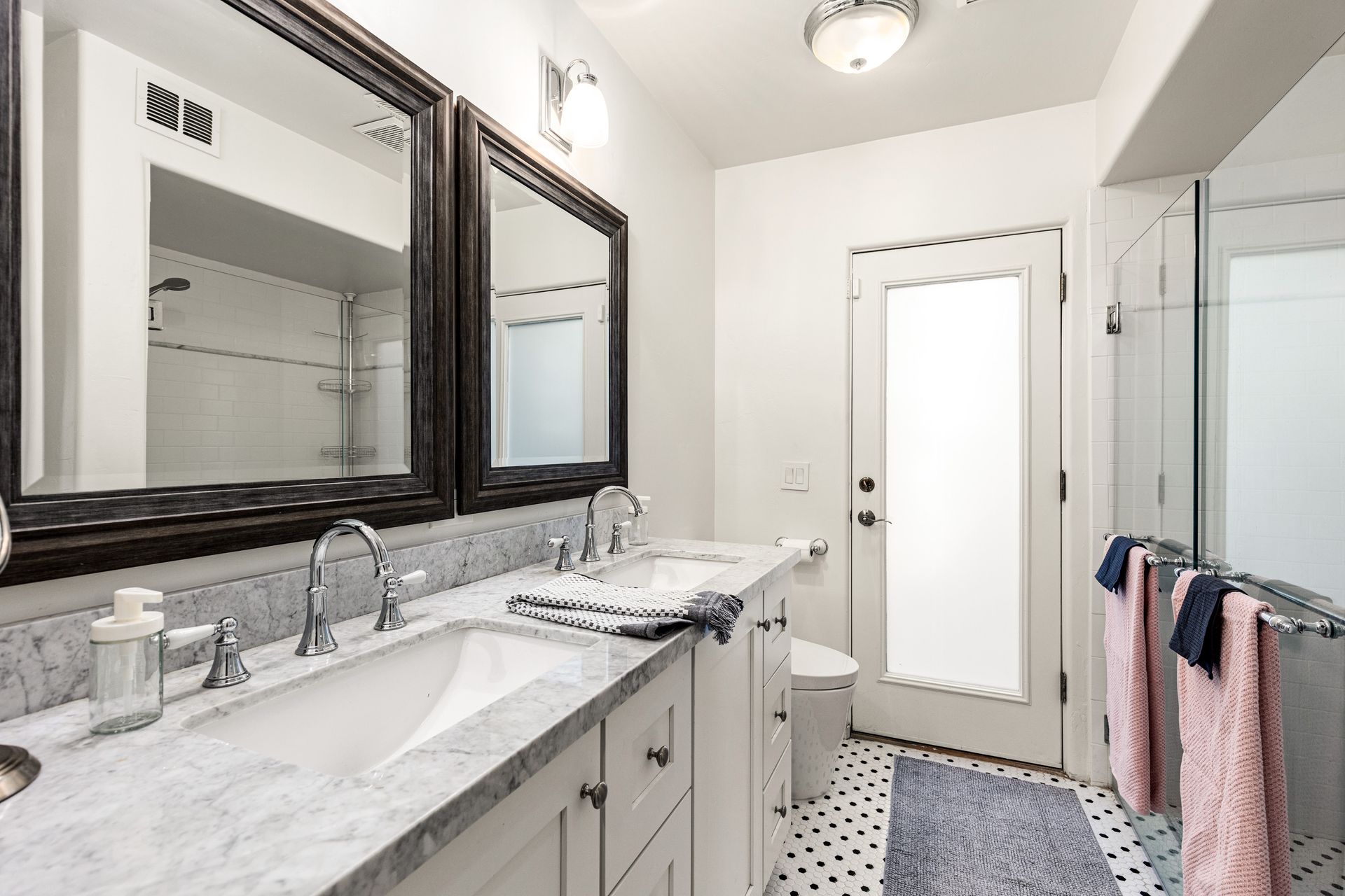 Bathroom with white cabinets, marble countertop, large mirrors, and a frosted glass door. Towels are pink and gray.