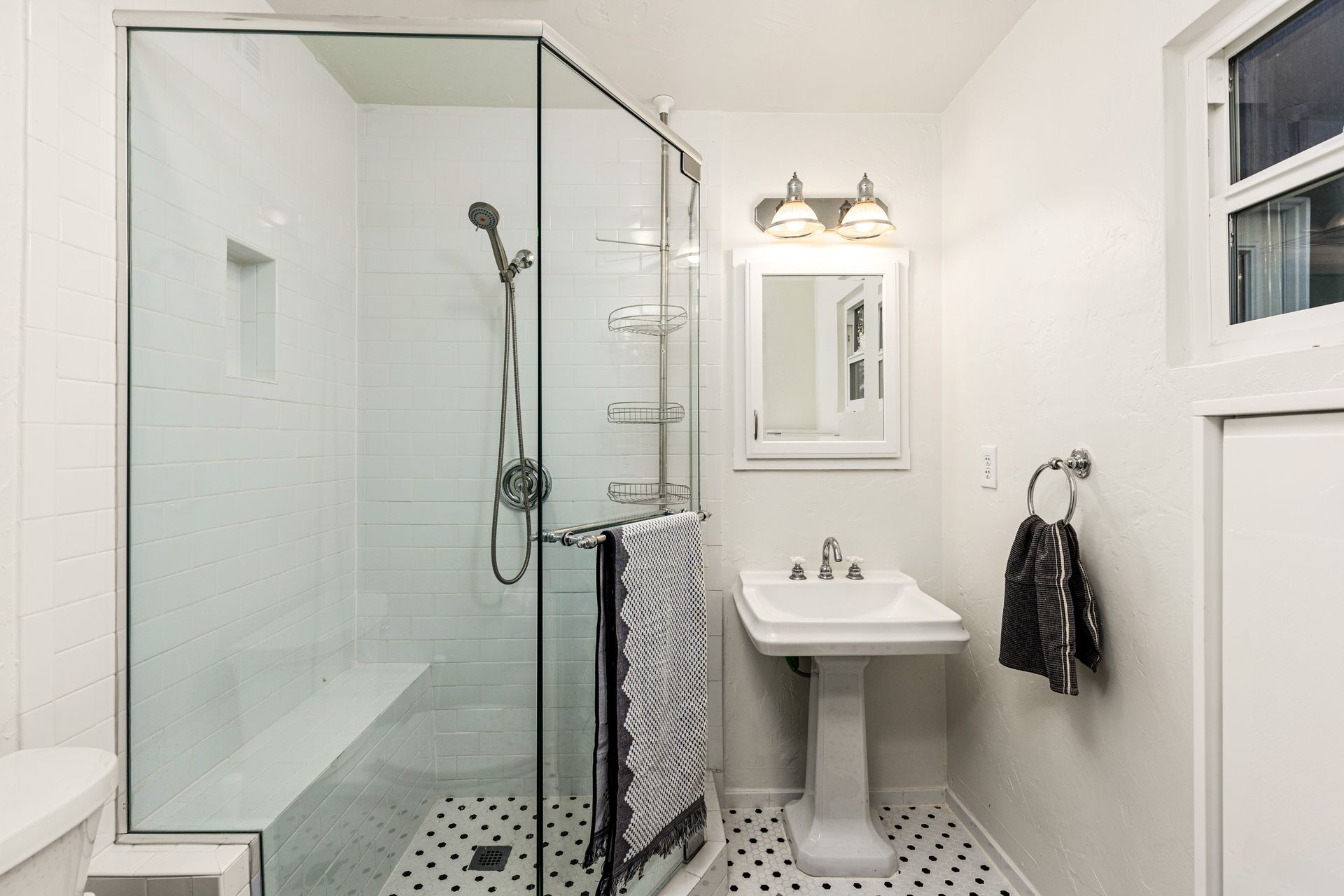 Small white bathroom with a glass shower, pedestal sink, and black and white tiled floor. A black and white towel hangs on a wall-mounted ring.
