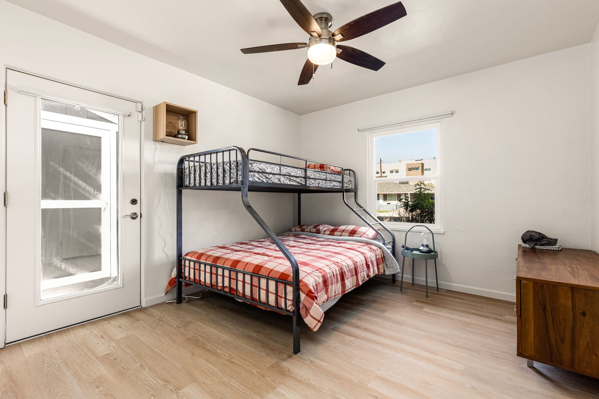 A bedroom with a bunk bed, a wooden dresser, and a door leading outside. The room has a window, a ceiling fan, and light-colored flooring.