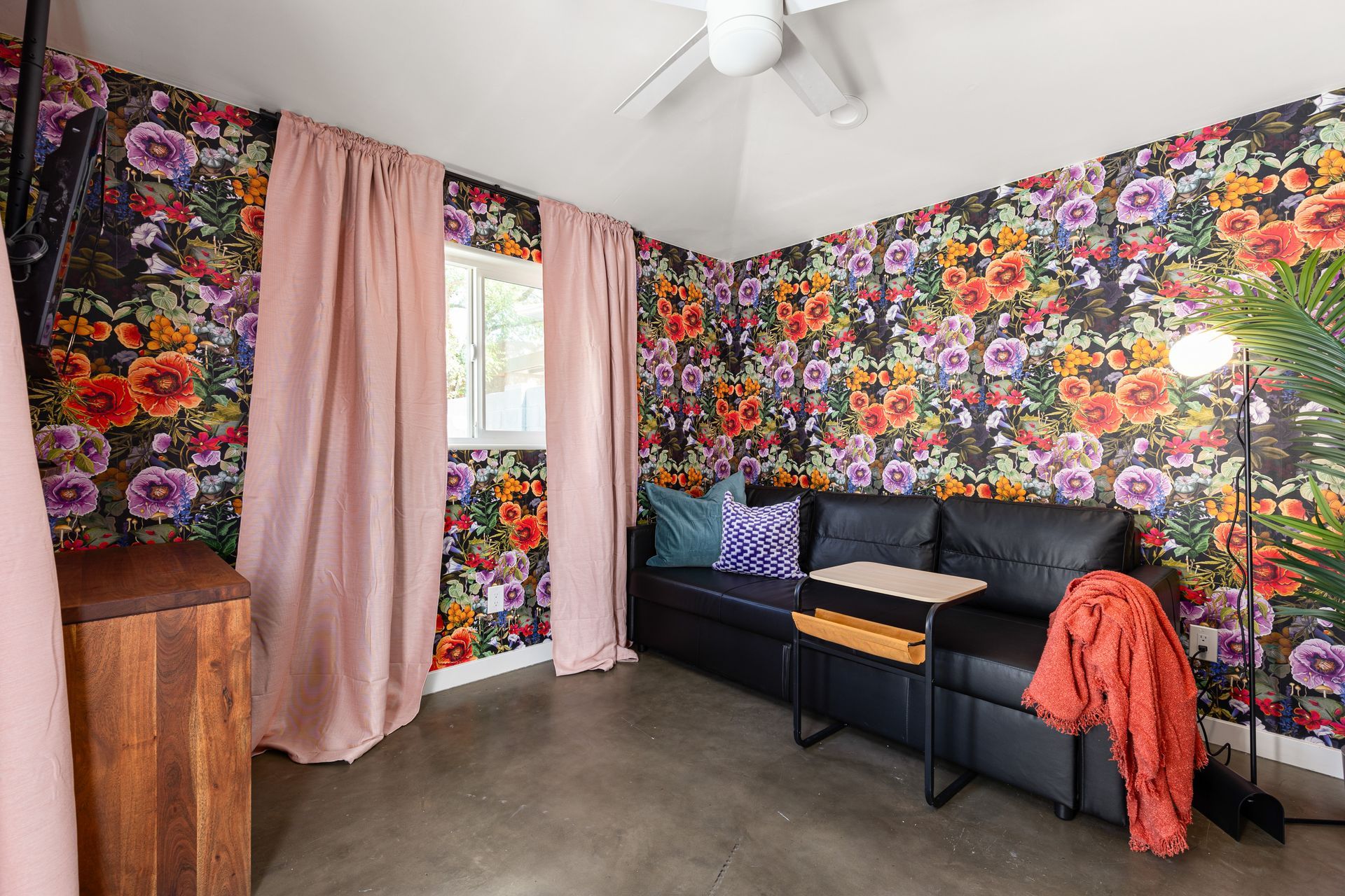 A small room with floral wallpaper, pink curtains, a black sofa, and a floor lamp.