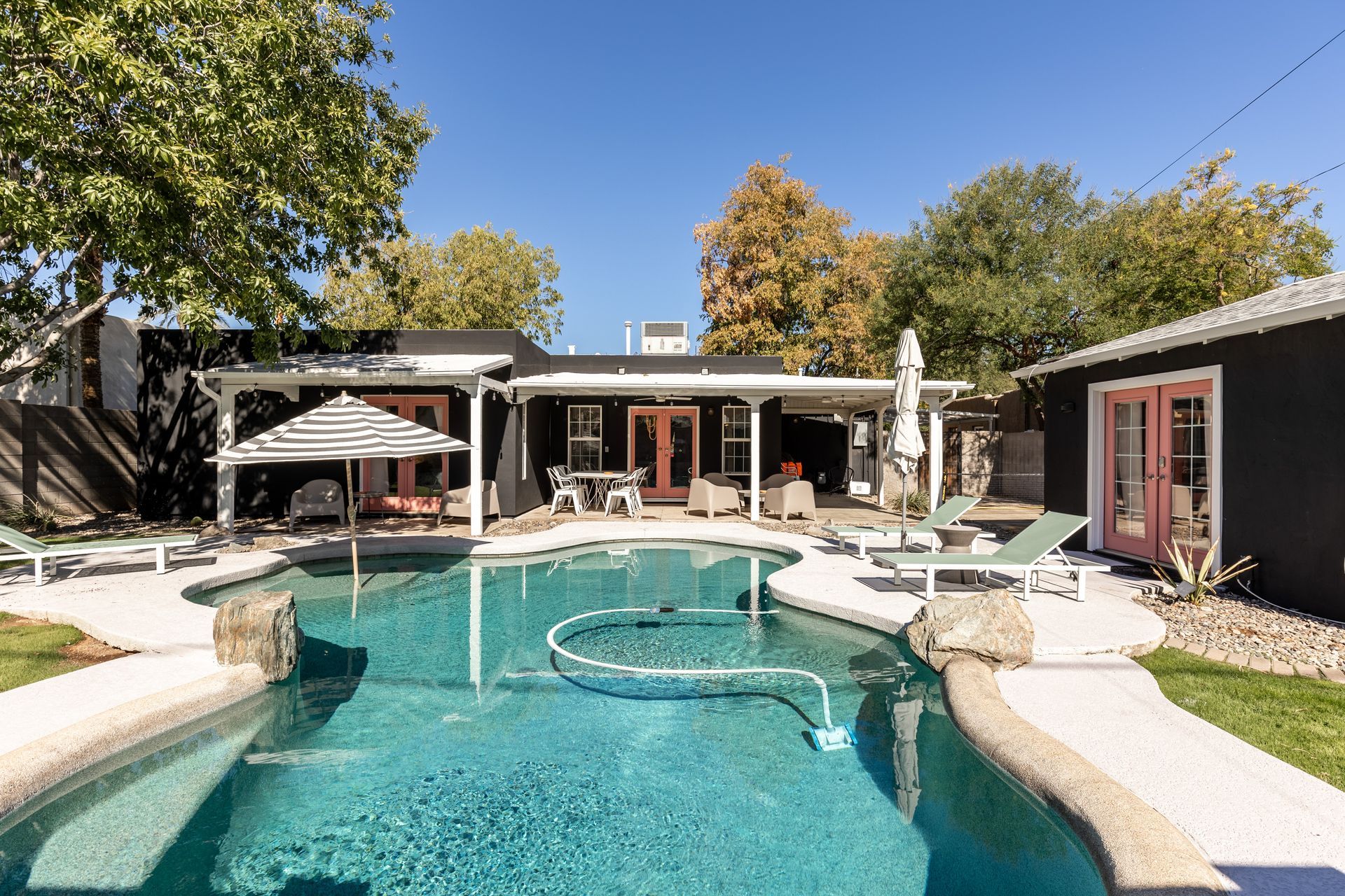 Backyard pool with lounge chairs, patio seating, and a black-painted house with red doors.