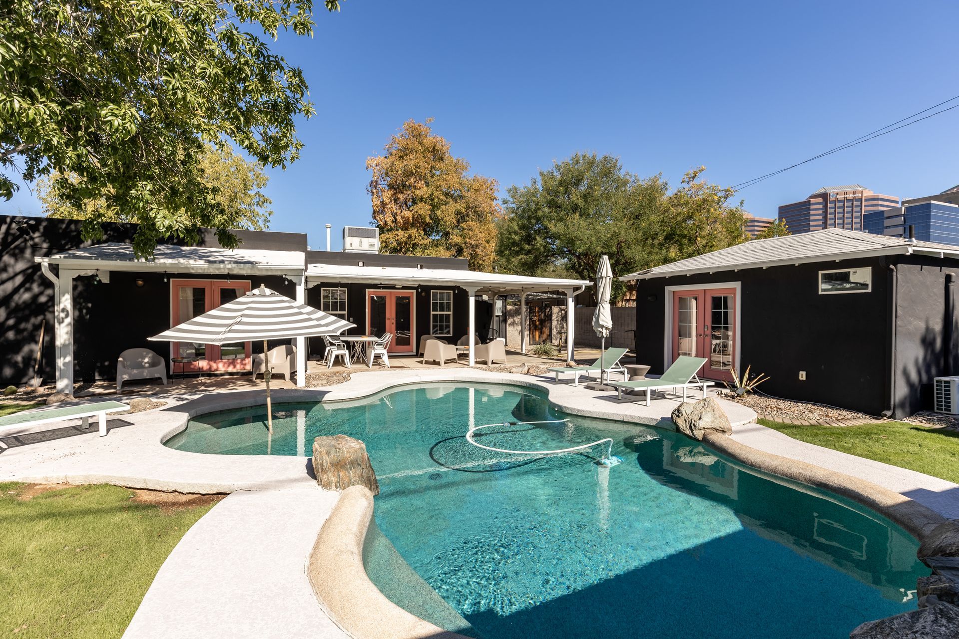 A backyard with a pool, patio, and two buildings, all with a modern design. The pool has light blue water, and the buildings have black siding with pink doors.