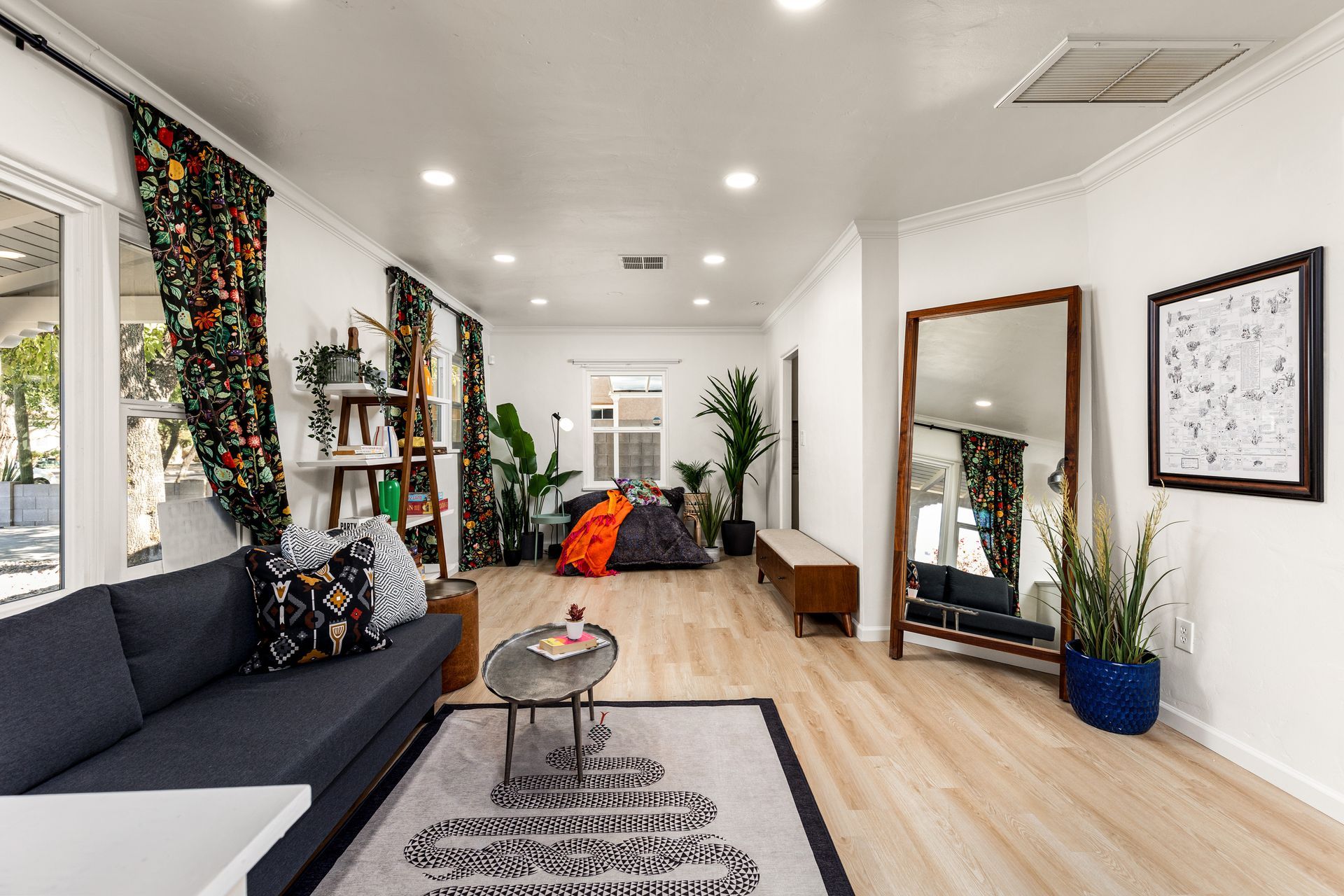 Bright living room with a dark blue sofa, large mirror, and patterned curtains. Plants and decorative items are placed throughout.