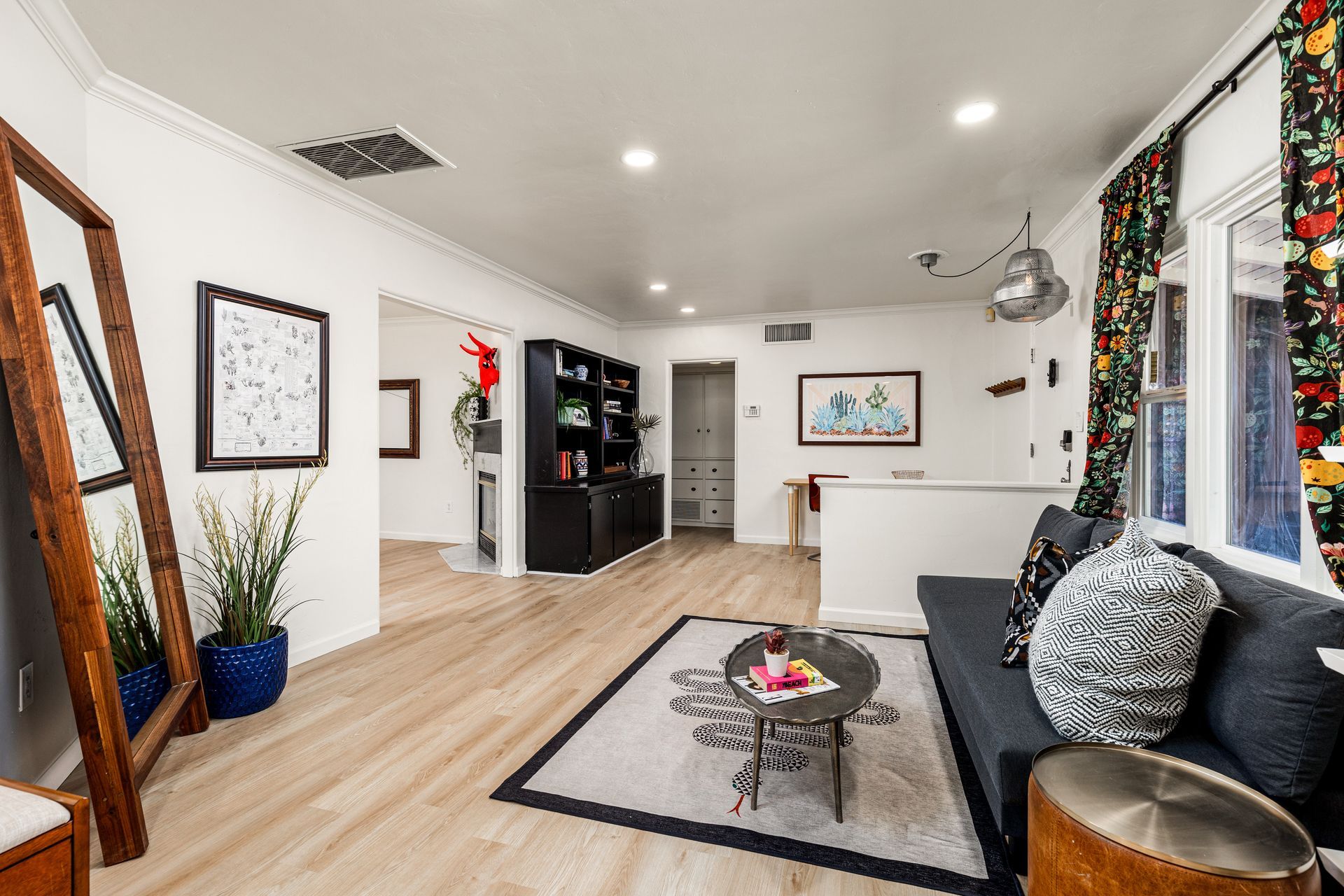 A modern living room with light wood floors, white walls, and a dark blue sofa with patterned pillows. A large mirror leans against a wall near a plant.