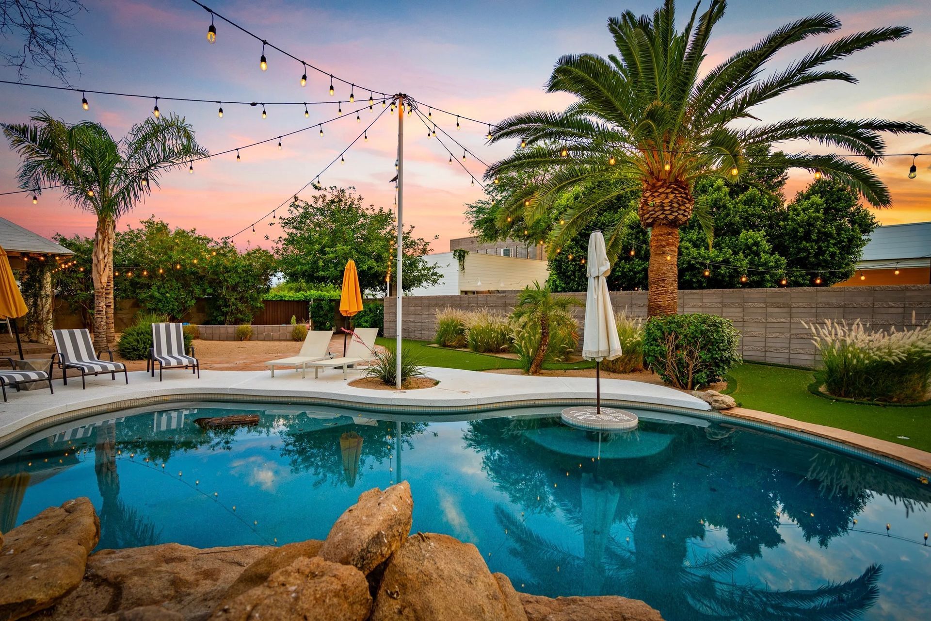 Backyard pool with string lights, palm trees, and a colorful sunset sky. Lounge chairs and umbrellas are arranged around the pool.