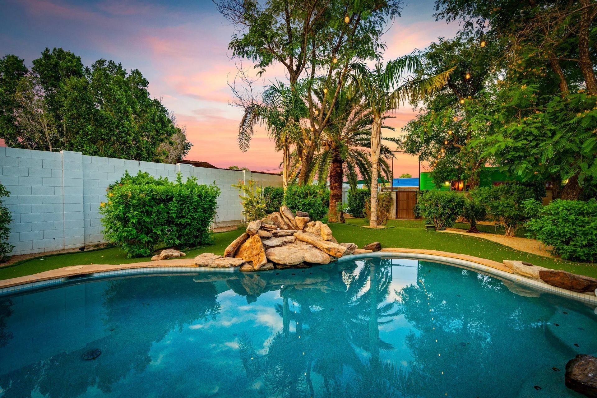 A backyard with a swimming pool, lush greenery, and a waterfall feature. The sky is a gradient of pink and blue.