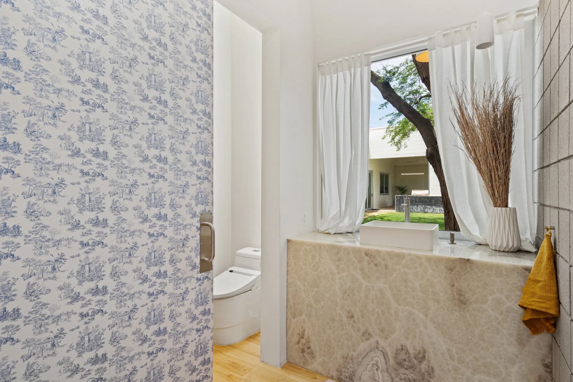 Bathroom interior with floral wallpaper, a white toilet, a stone sink, and a window with white curtains.