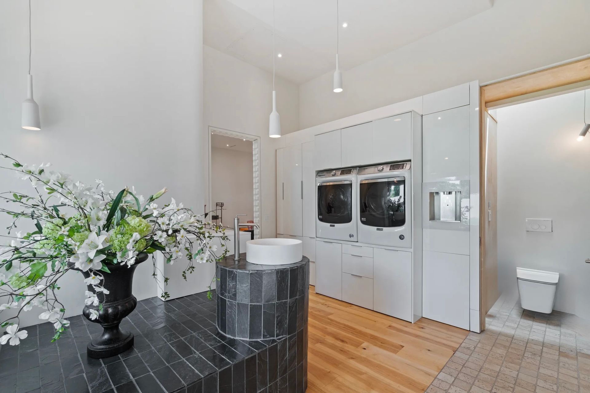 A bright, modern laundry room with white walls, wooden floors, and a black and gray tiled counter with a floral arrangement. A washer and dryer are built-in on the right.