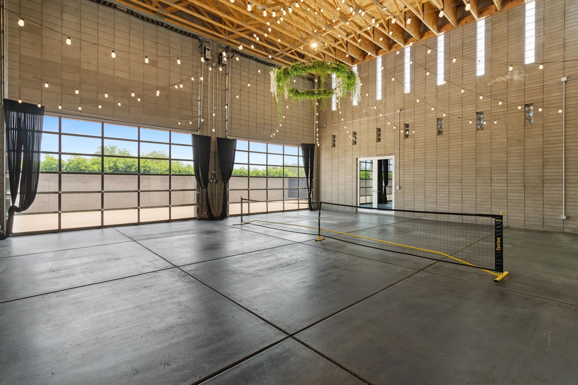 Indoor event space with a pickleball net set up, open glass doors to outside, string lights.