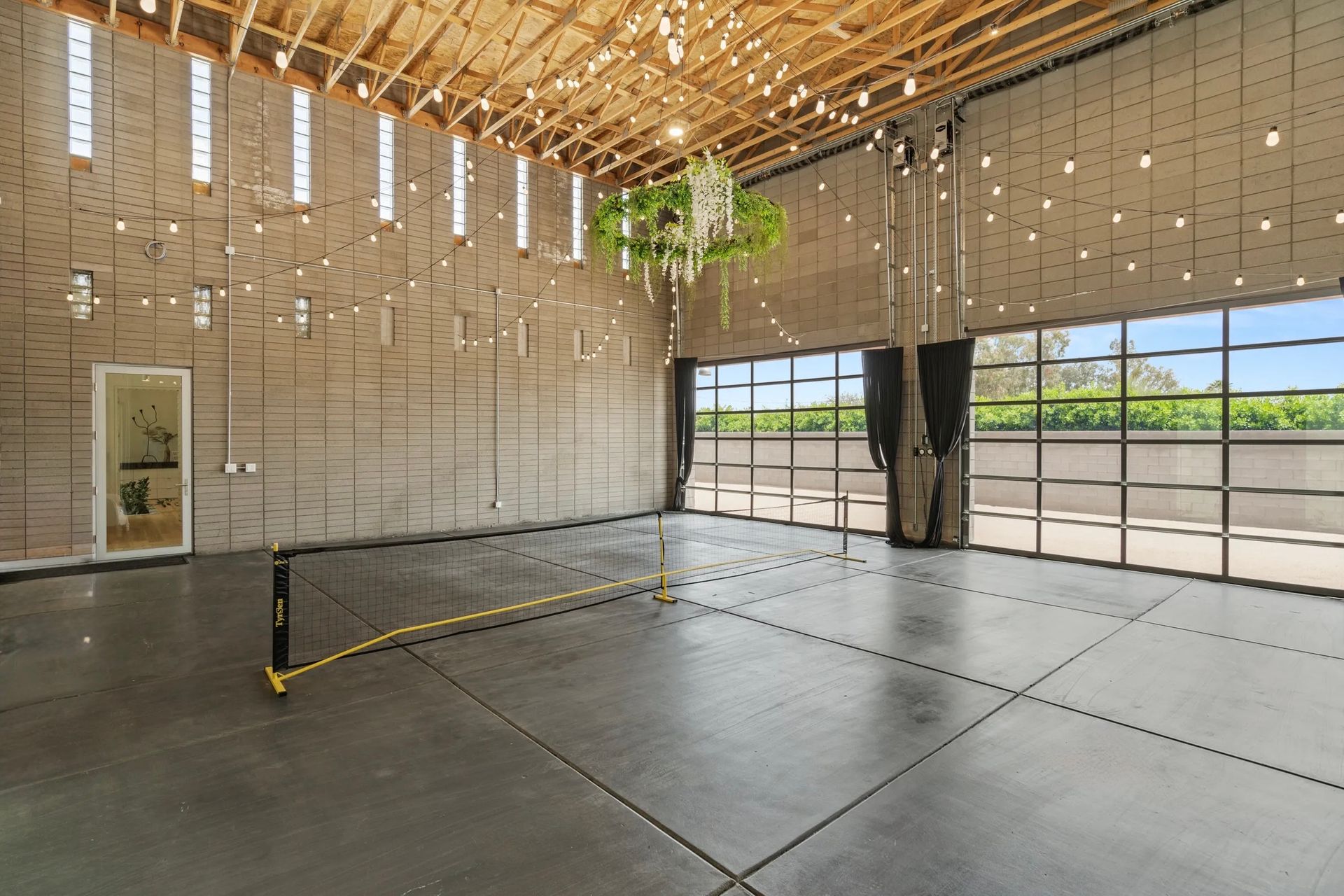 Empty event space with a concrete floor, large windows, and string lights hanging from the ceiling. A volleyball net is set up.