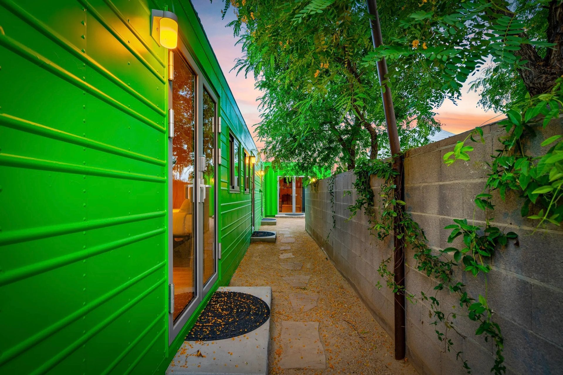A narrow walkway between a bright green building and a gray cinder block wall, with foliage and a setting sun.