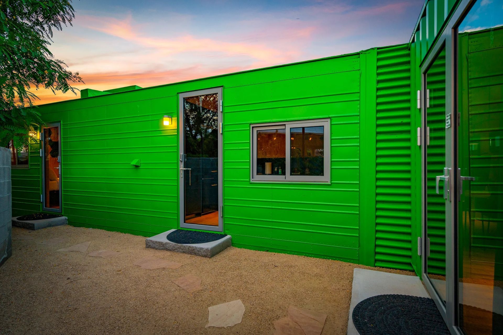 Bright green corrugated metal building exterior with doors, windows, and gravel ground, under a colorful dusk sky.