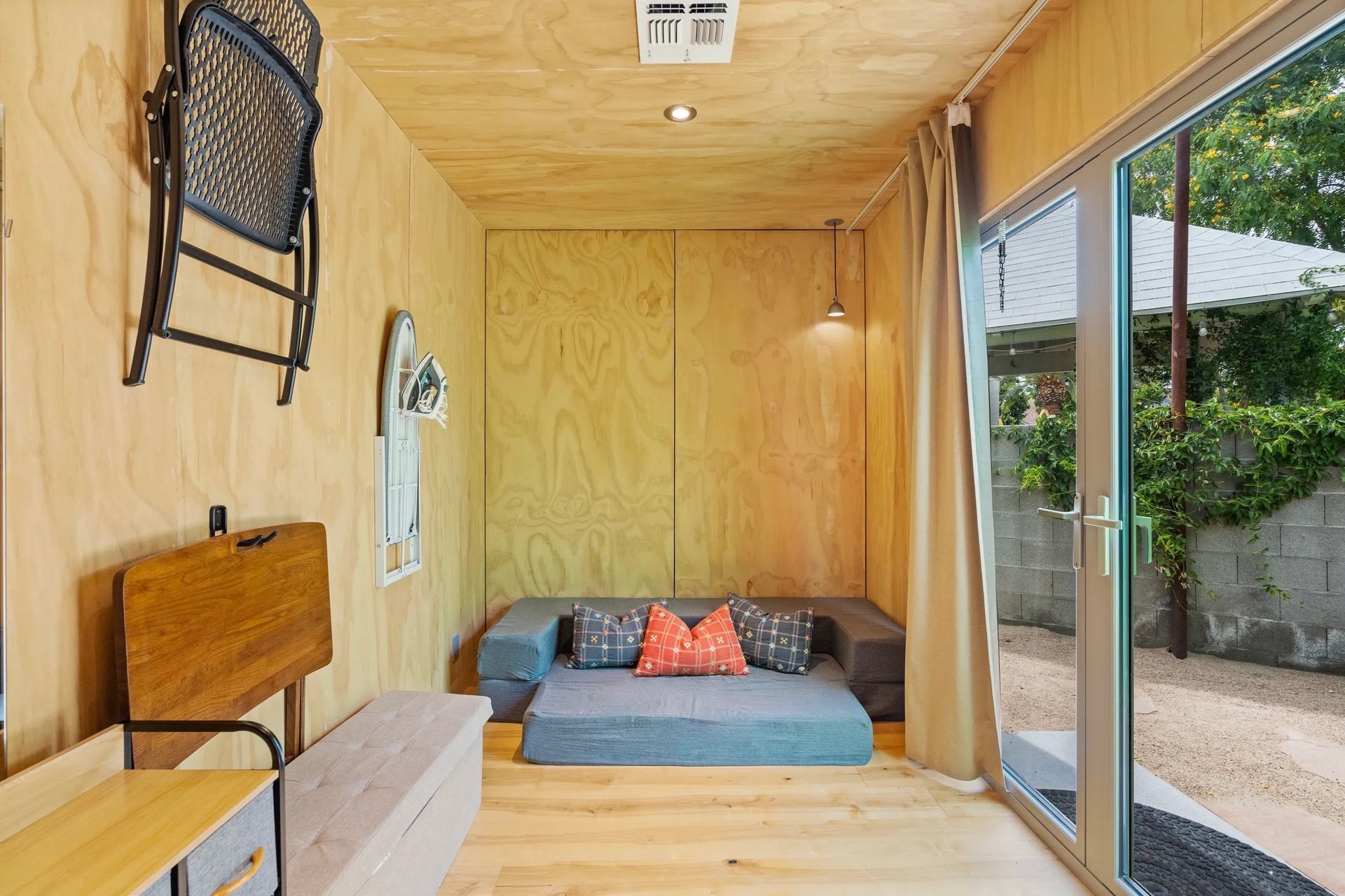 Interior of a small, modern room with plywood walls, a gray sofa, and sliding glass doors leading to a yard.