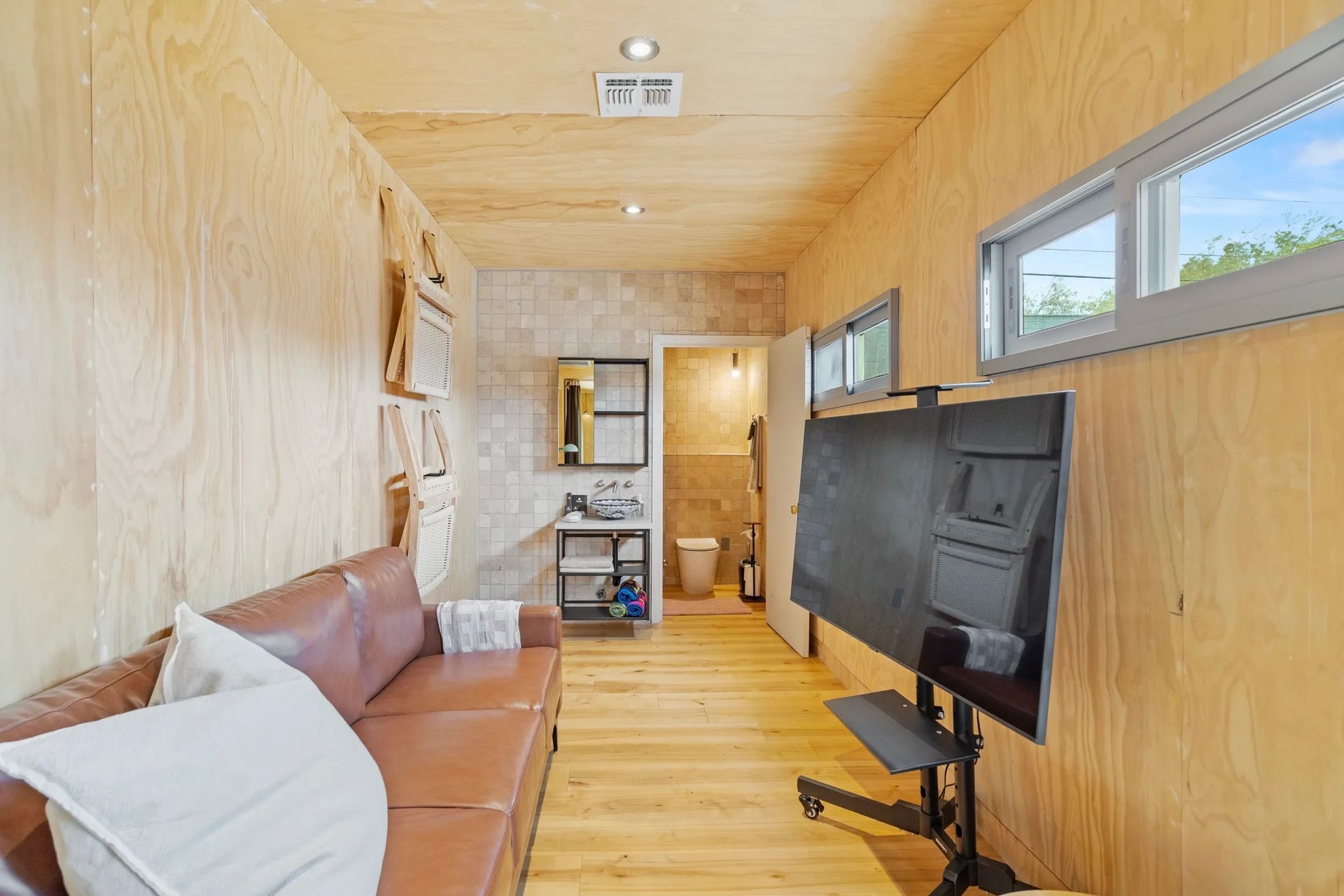 A small, modern room with wood paneling, a brown leather couch, and a television on a stand, with a bathroom visible at the end.