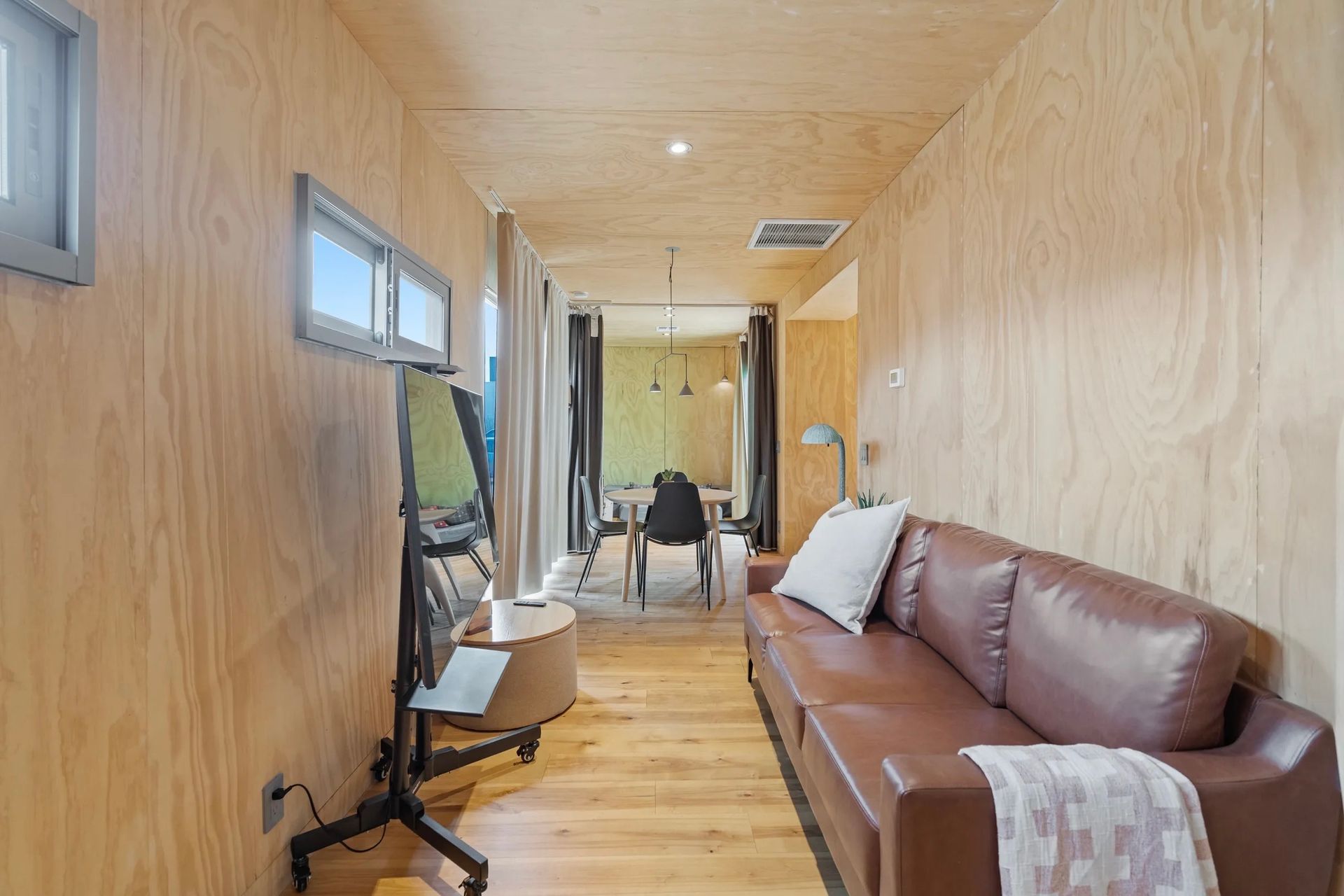 A narrow living space with wood-paneled walls, a brown leather sofa, and a TV on a stand. Windows provide natural light.