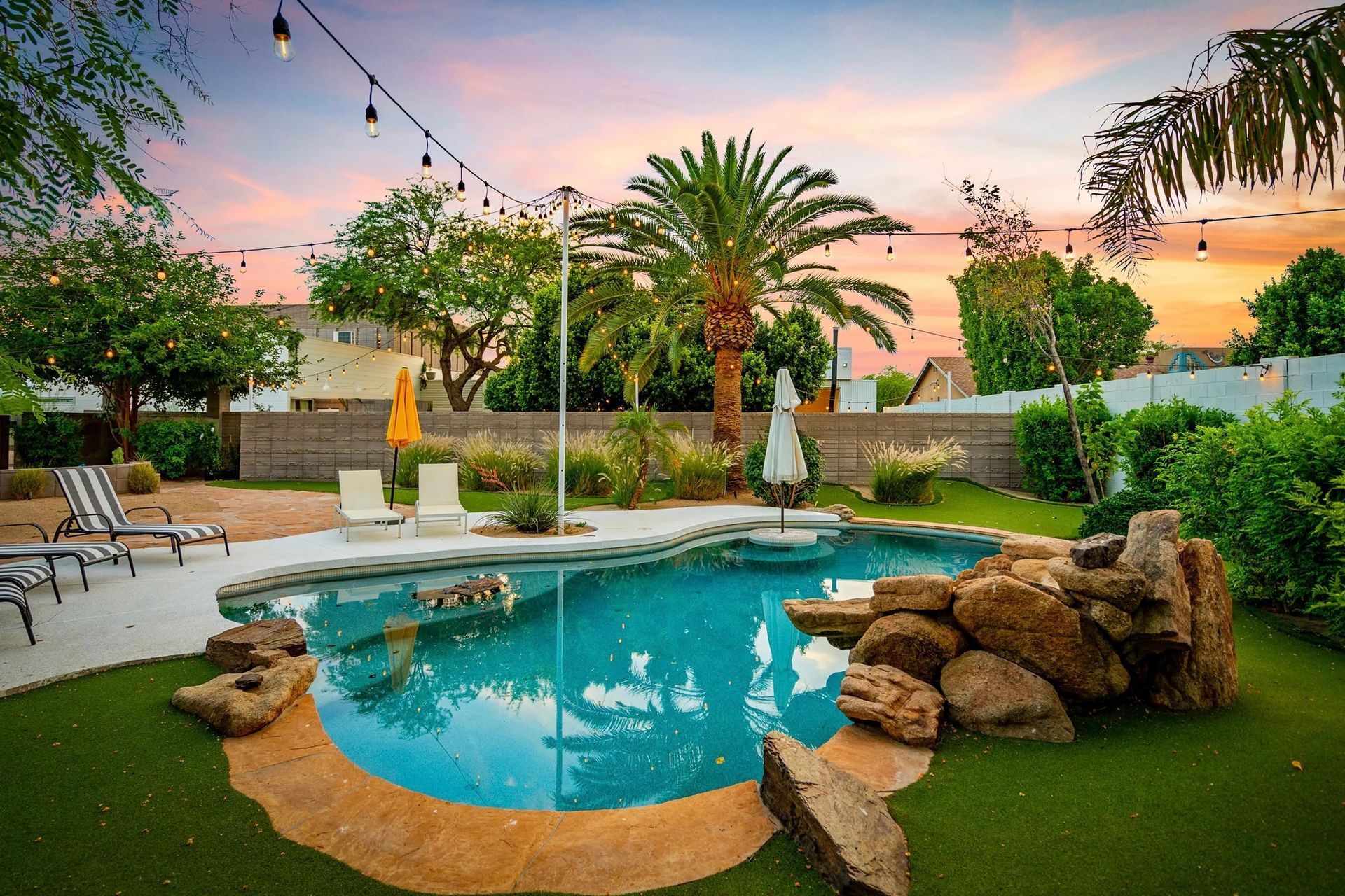 Backyard pool with rocks, artificial turf, and a palm tree under a colorful sunset sky. String lights are strung overhead.