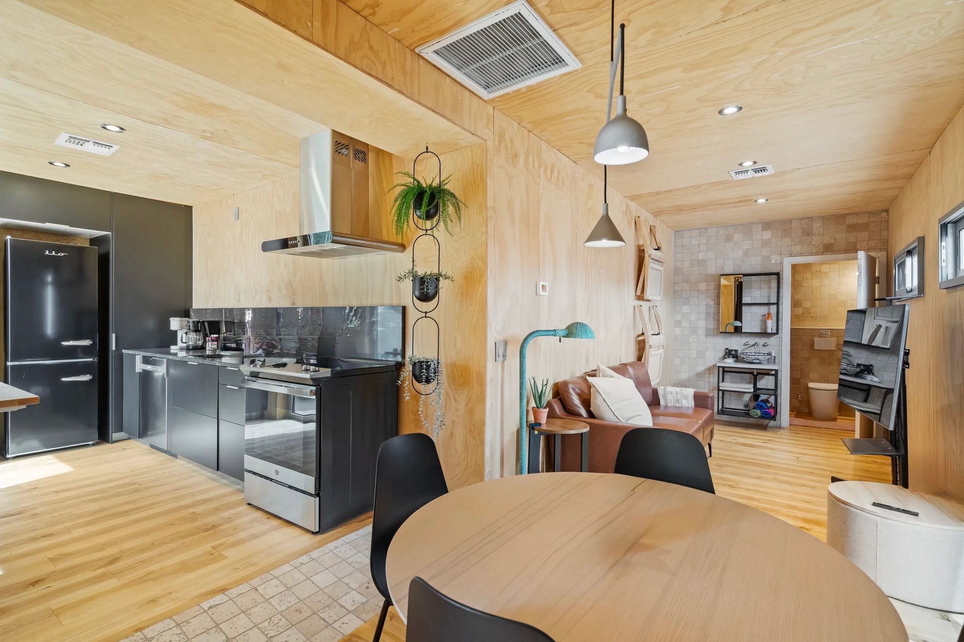 Interior view of a modern apartment with wood paneling. The kitchen features black appliances, while a living area has a brown sofa, and round table.