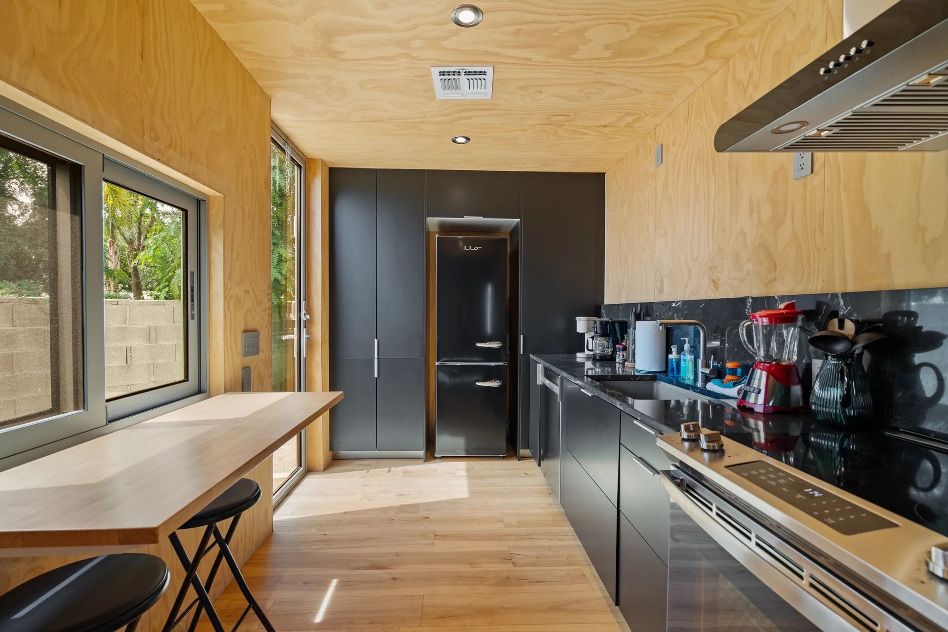 Modern kitchen with black cabinetry, appliances, and countertops, contrasted by light wood paneling and a window with a built-in counter.