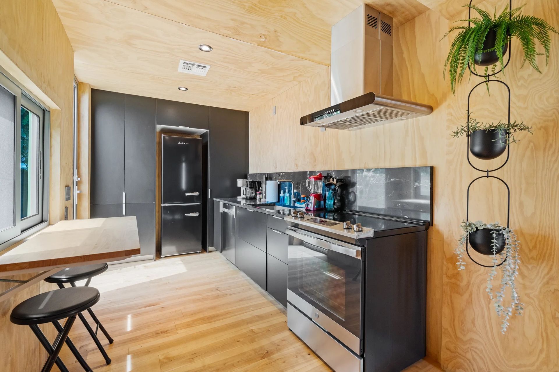 A modern kitchen with wooden walls and floor. It features dark gray cabinets, stainless steel appliances, and hanging plants.