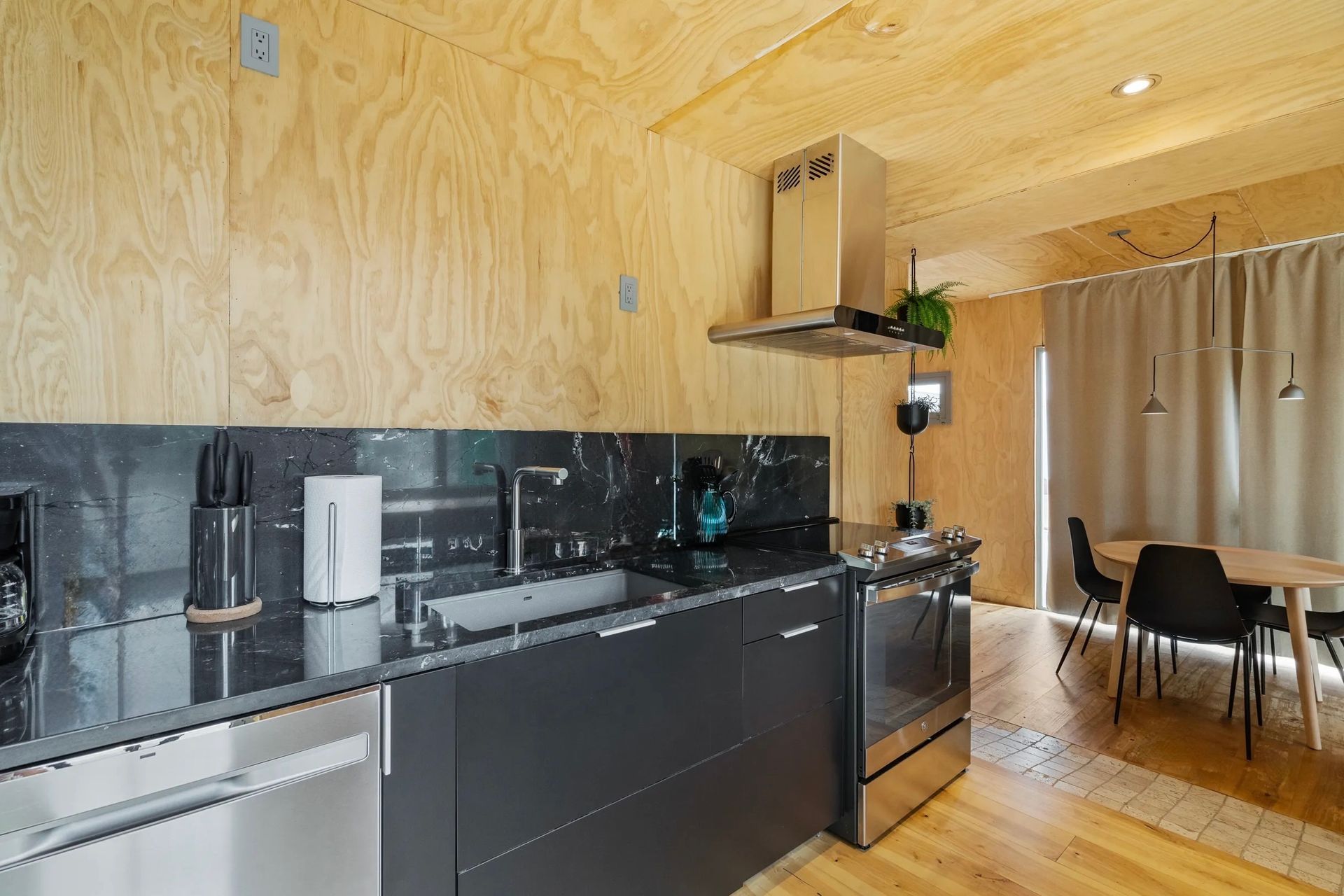 Modern kitchen with wooden walls and ceiling, gray countertops, black backsplash, and stainless steel appliances. Dining table with chairs in the background.