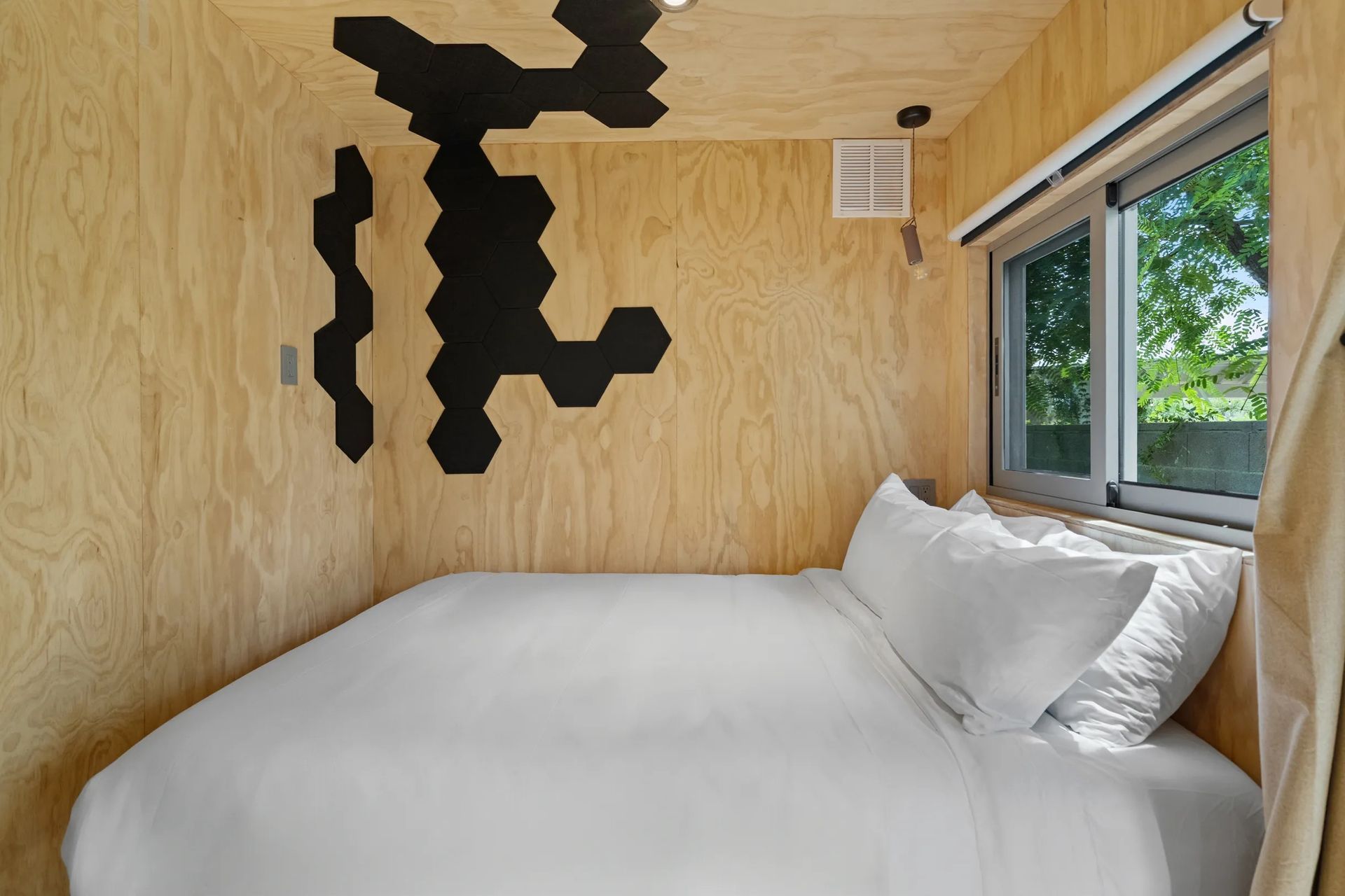 Small bedroom with a bed, a window, and plywood walls with black hexagon art.