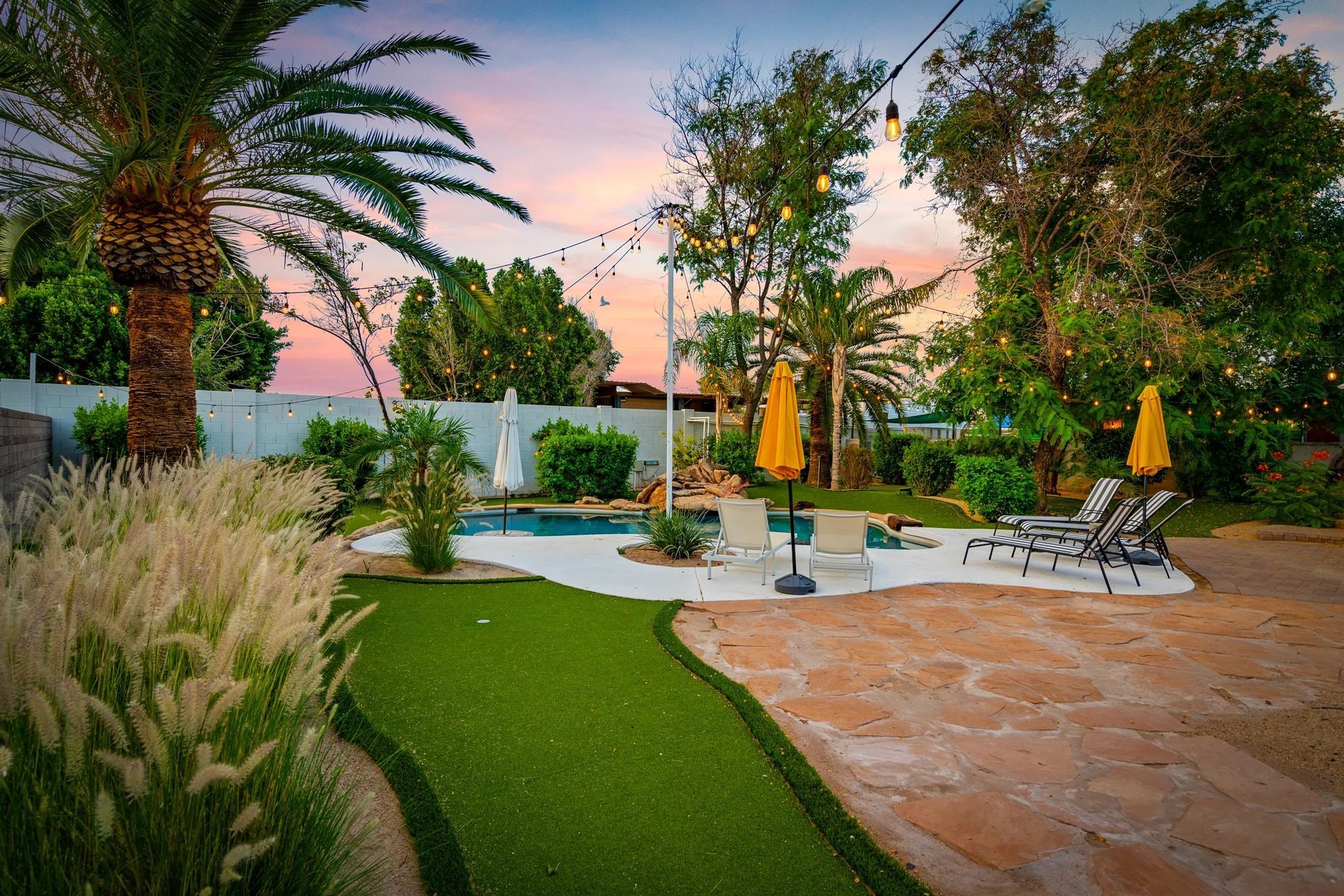 Backyard oasis with a pool, lounge chairs, yellow umbrellas, and string lights against a colorful sunset sky.