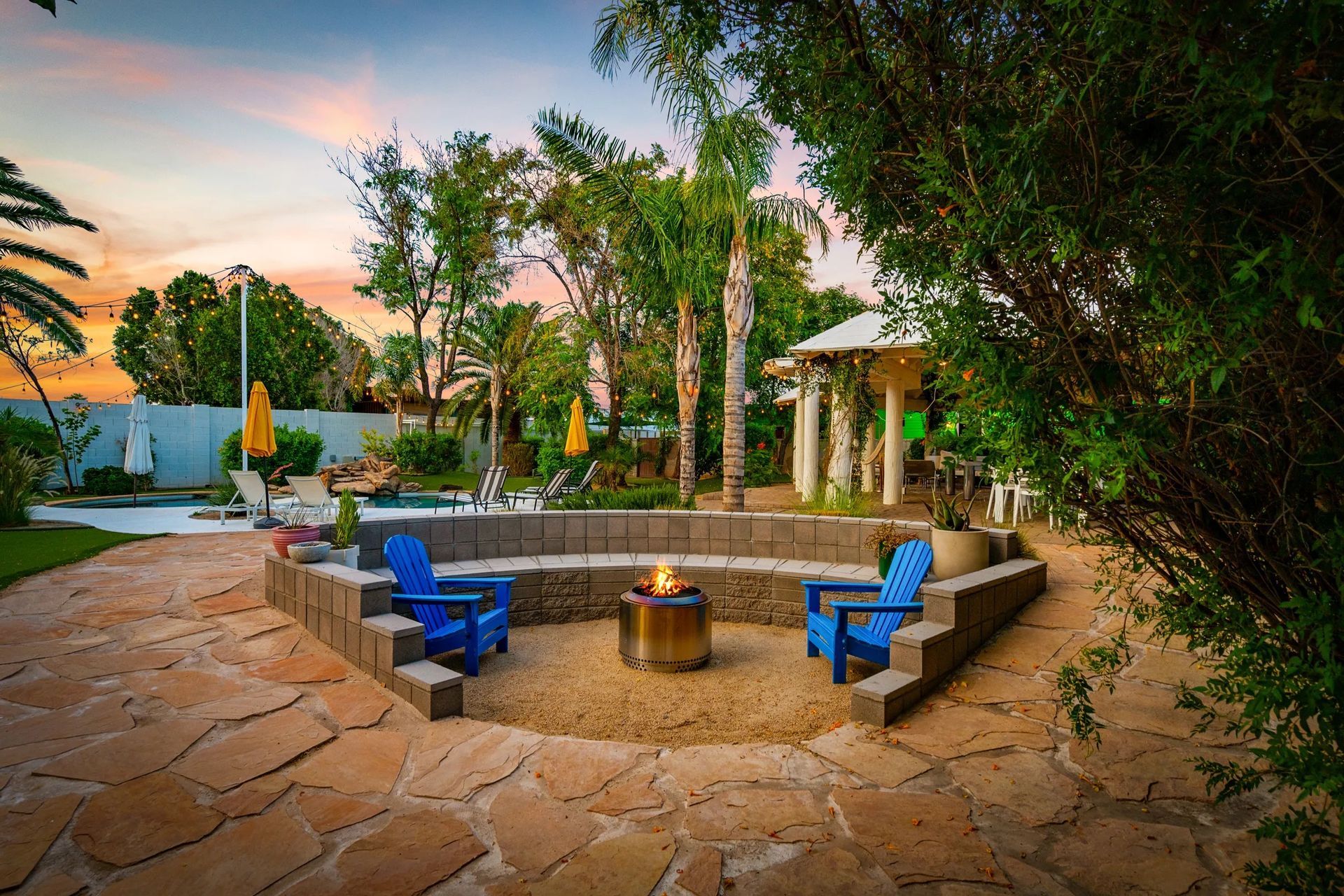 A backyard patio with a fire pit, blue chairs, and a gazebo. The scene is lit by an evening sunset.