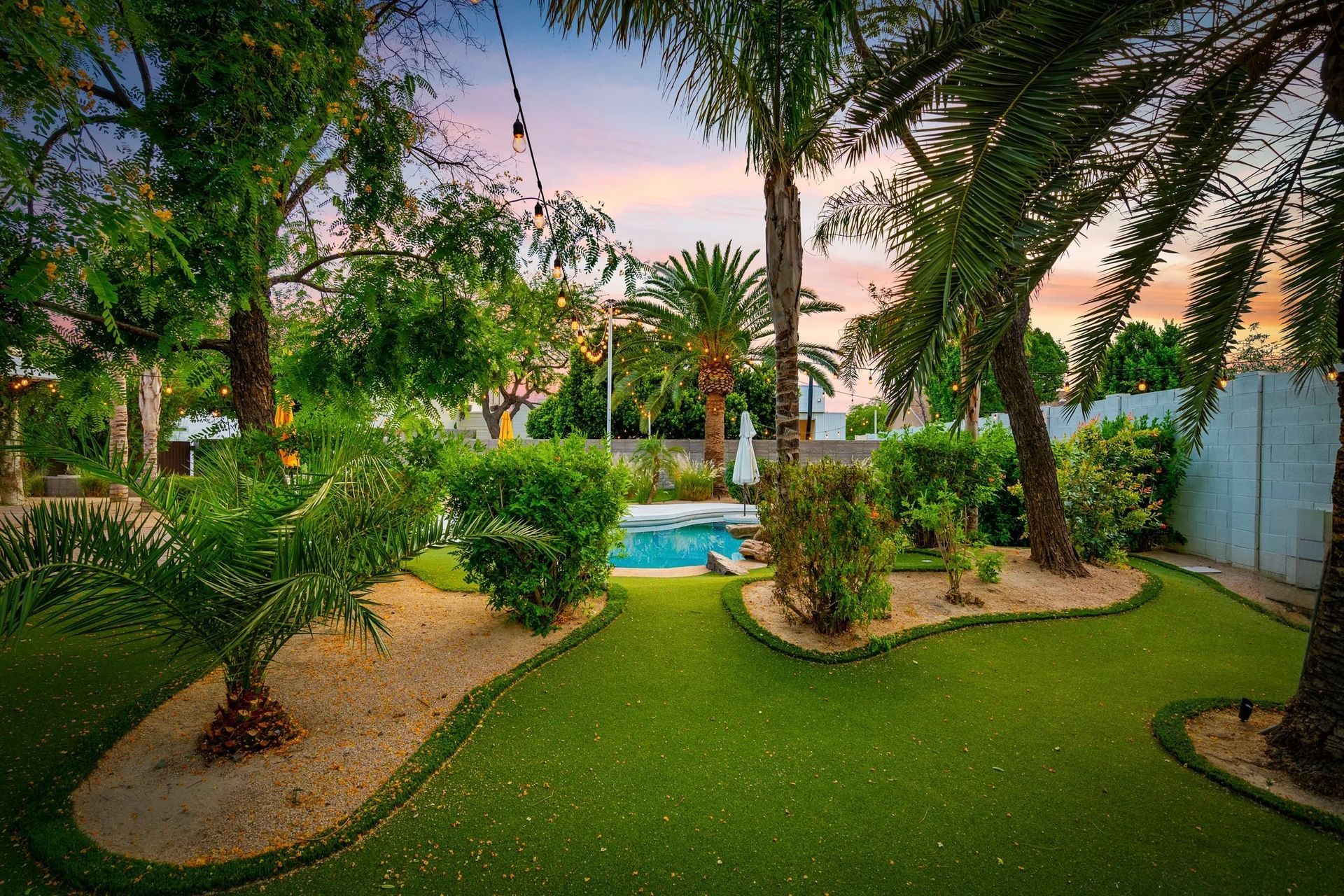 Lush backyard with a small pool, surrounded by palm trees, green lawn, and flowering bushes against a colorful sunset.