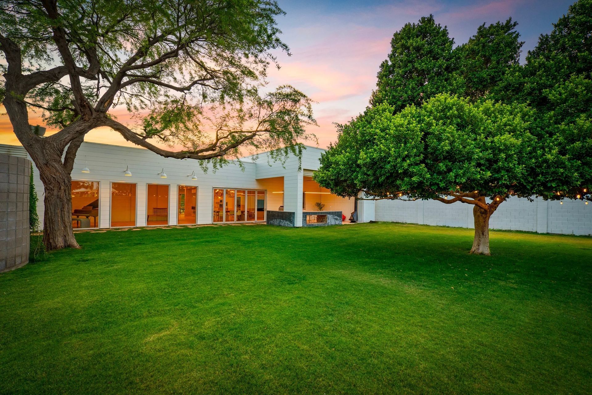 Backyard with lush green lawn, white building, and two large trees under a colorful sunset sky.