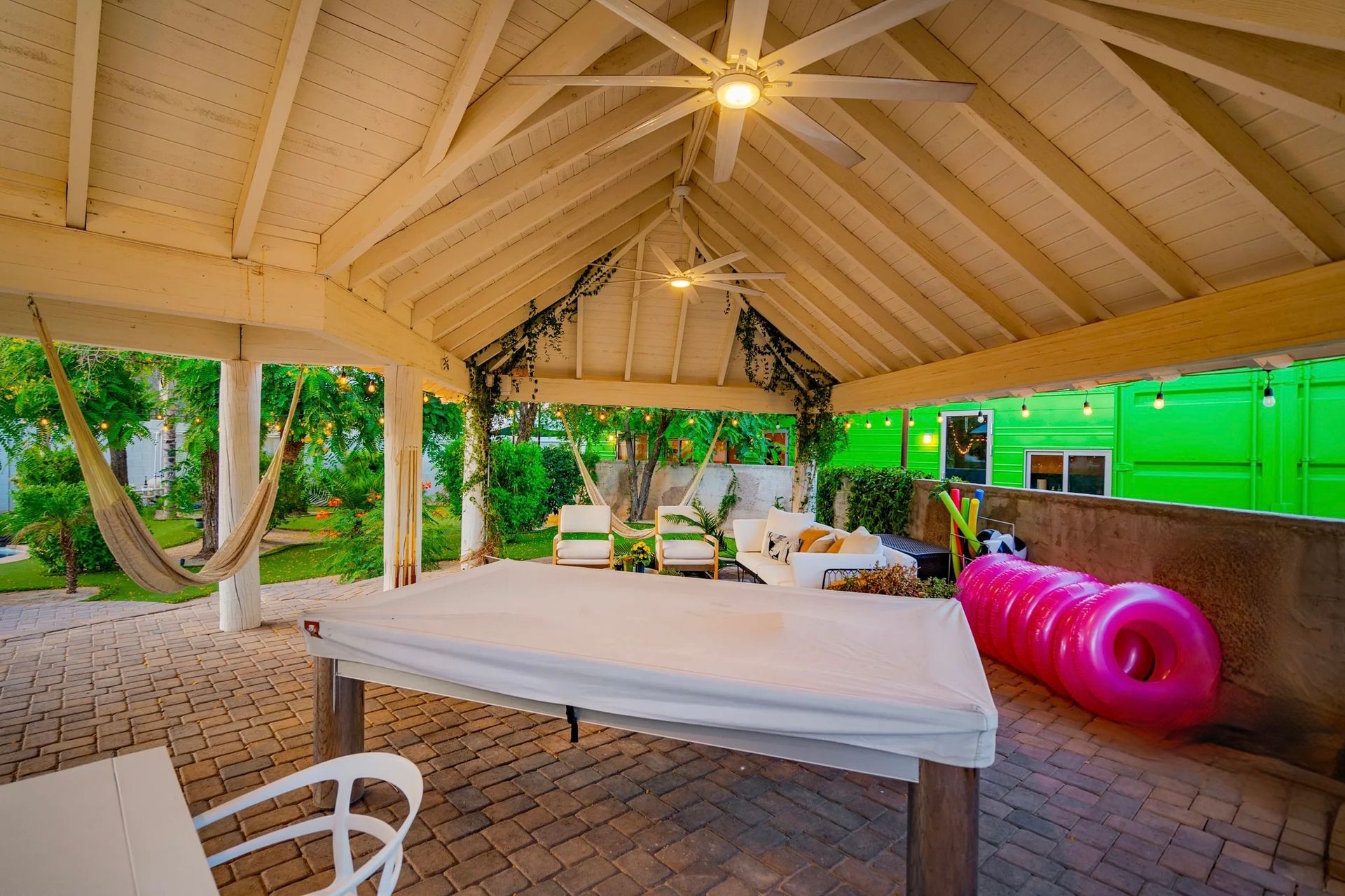 A covered patio with a table, hammock, and seating. The space features a white ceiling, green walls, and a pink inflatable.