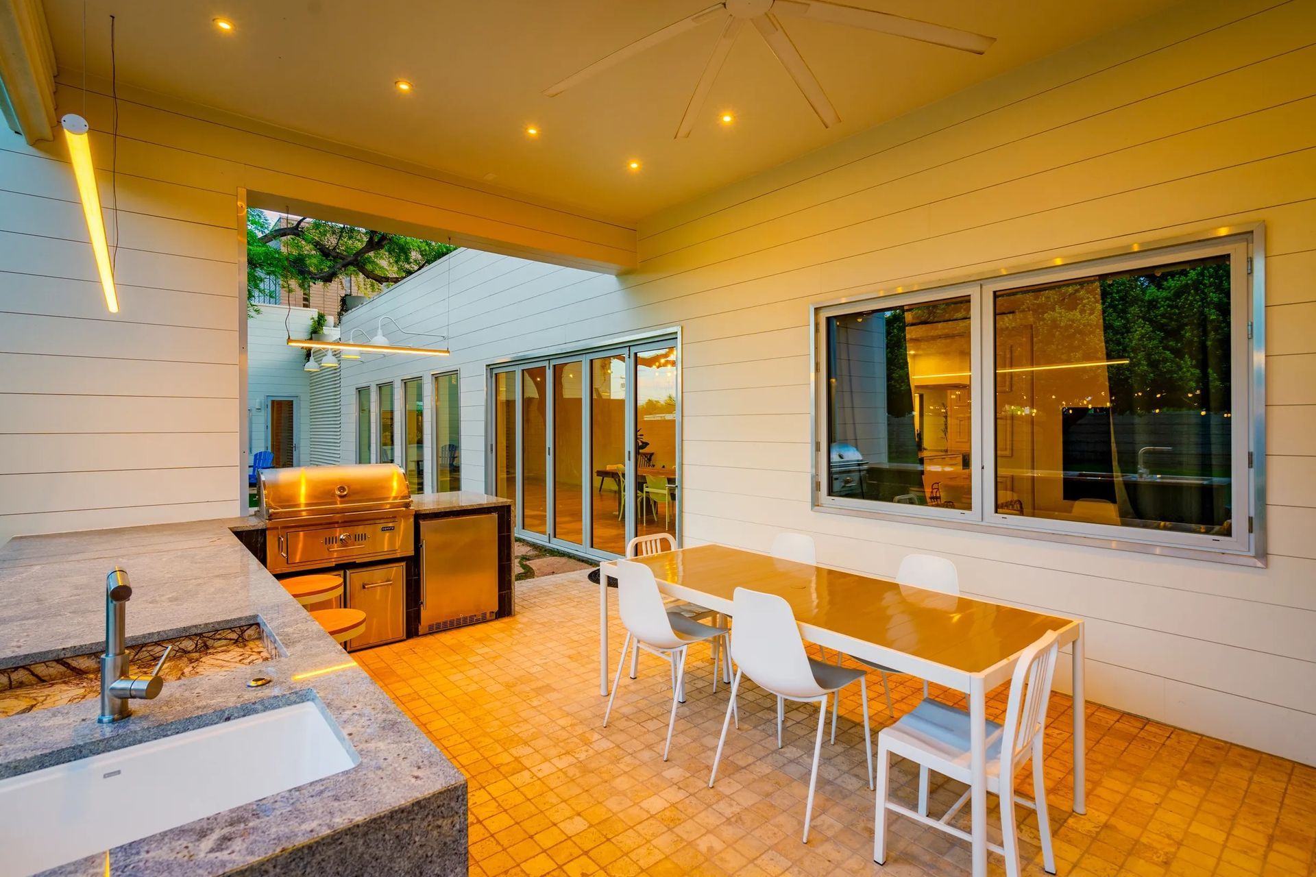 Outdoor kitchen with a long table, white chairs, and a grill area with stainless steel appliances. The space has a warm color palette.