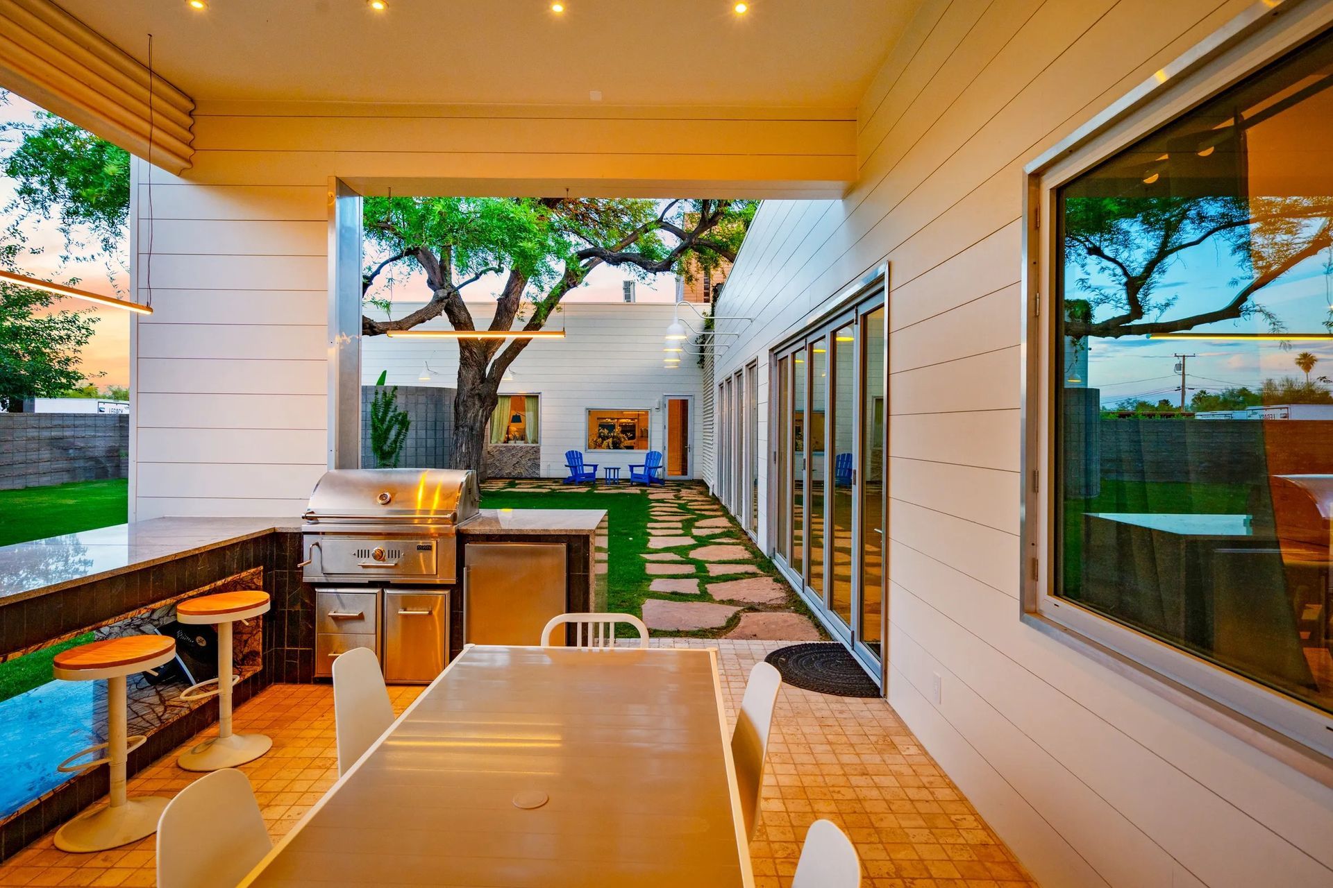 Outdoor patio with a grill, dining table, and a view of a backyard with a tree, blue chairs, and a pathway.
