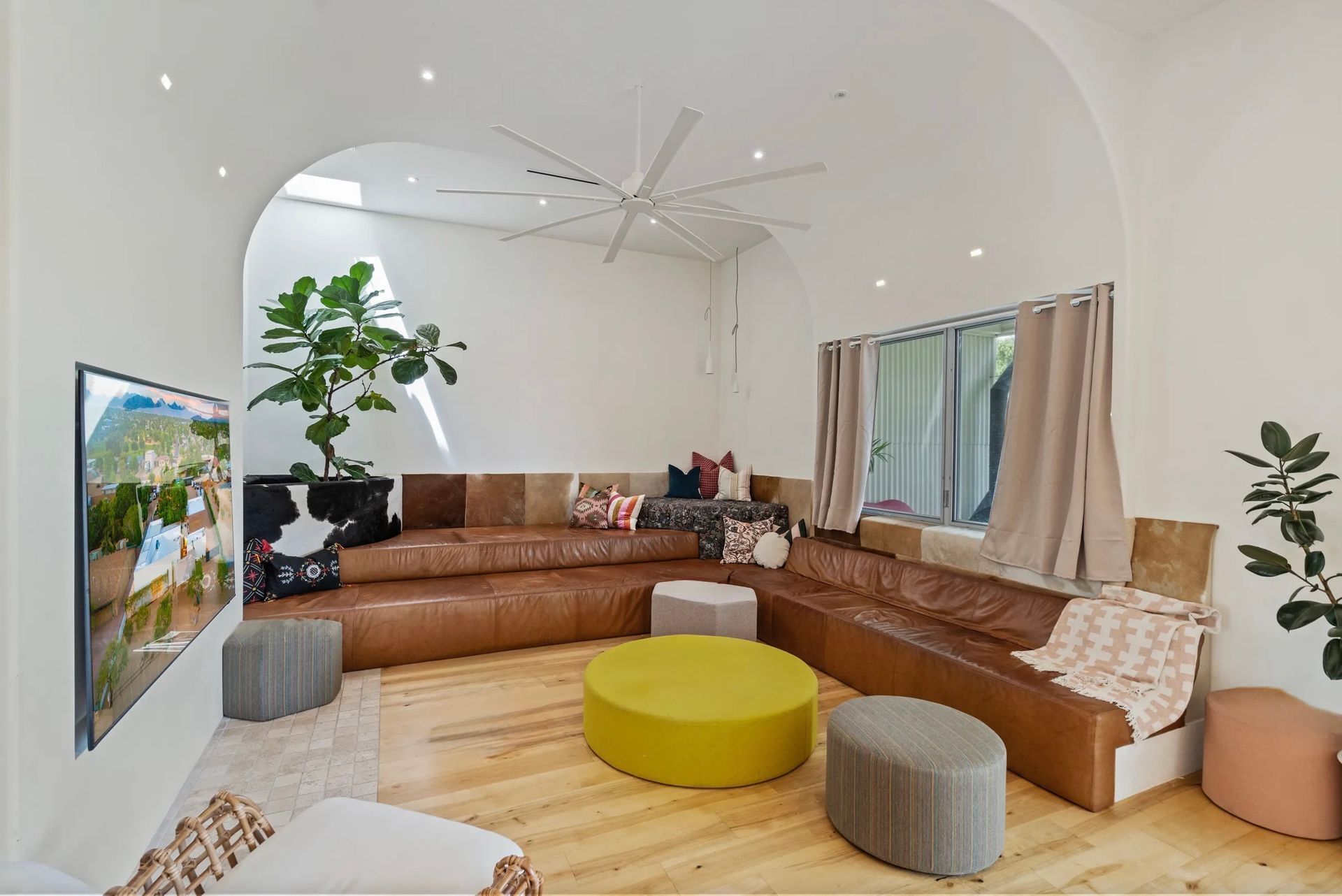 Bright, modern living room with a brown sectional sofa, various ottomans, and a large plant in a neutral setting.