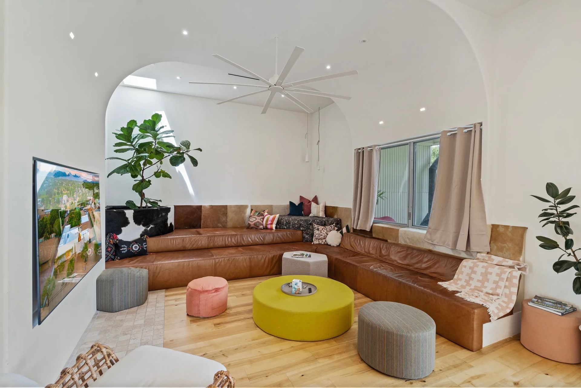 A modern living room with brown leather seating, colorful ottomans, and an archway leading to a plant.